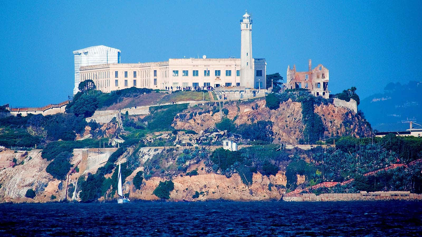 California Island 桌布 壁紙 — 一座歷史悠久的監獄堡壘，坐落在崎嶇的懸崖上，俯瞰深藍色的海水，一艘帆船從下方駛過。