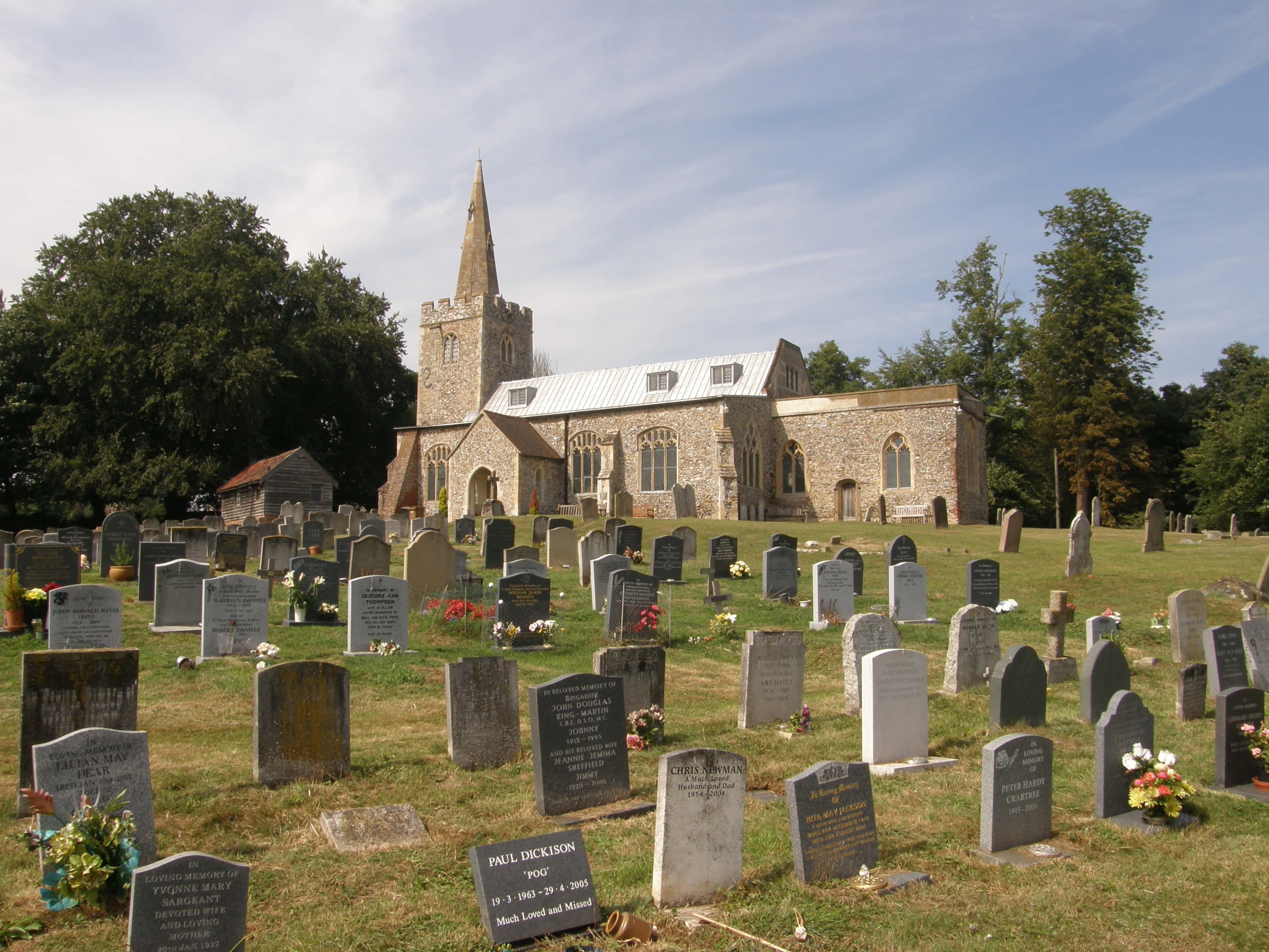 Gravestone 桌布 壁紙 — 一座歷史悠久的石砌教堂，擁有高聳的尖頂，矗立在陽光普照的墓園後方，墓園中佈滿風化的墓碑和色彩鮮豔的花卉祭品。