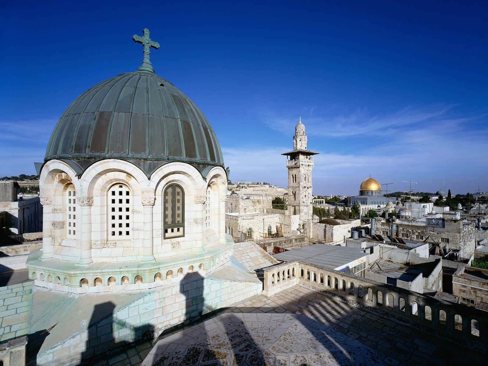 Israel 桌布 壁紙 — 一座歷史悠久的石頭教堂，擁有綠色銅圓頂和十字架，俯瞰著陽光明媚的城市景觀，遠處有一座金色圓頂清真寺，在清澈的藍天下。