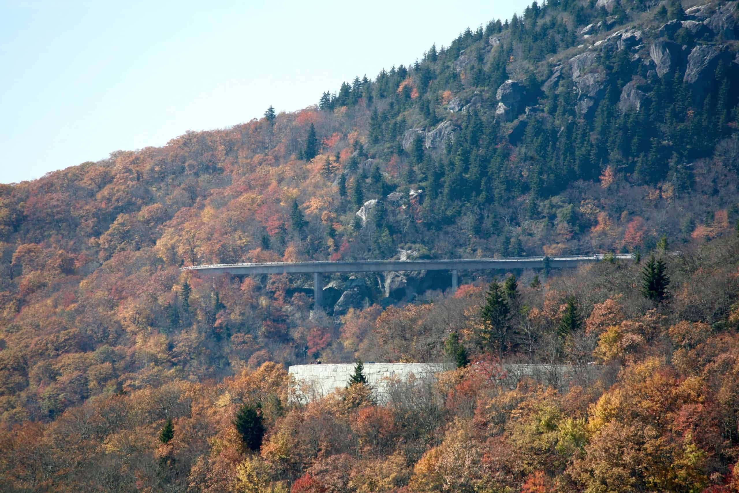 Blue Ridge Parkway 桌布 壁紙 — 一座混凝土橋樑橫跨在岩石山脈上，山上覆蓋著秋葉，天空晴朗。