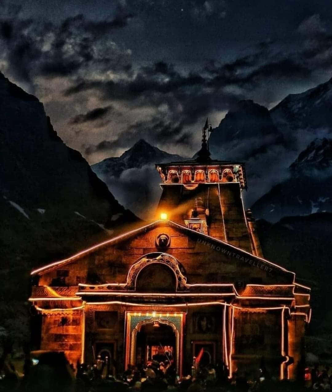 Kedarnath 桌布 壁紙 — 一座溫暖照明的寺廟在戲劇性的山區夜空下發光，背景有烏雲和遠峰。