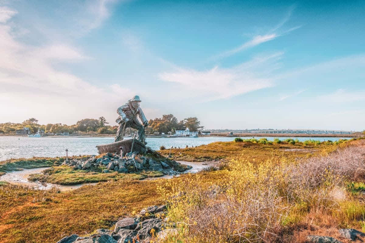 California Island 桌布 壁紙 — 一座漁夫在船上的銅像矗立在岩石島上，周圍是水和秋天的草，在明亮的藍天下。