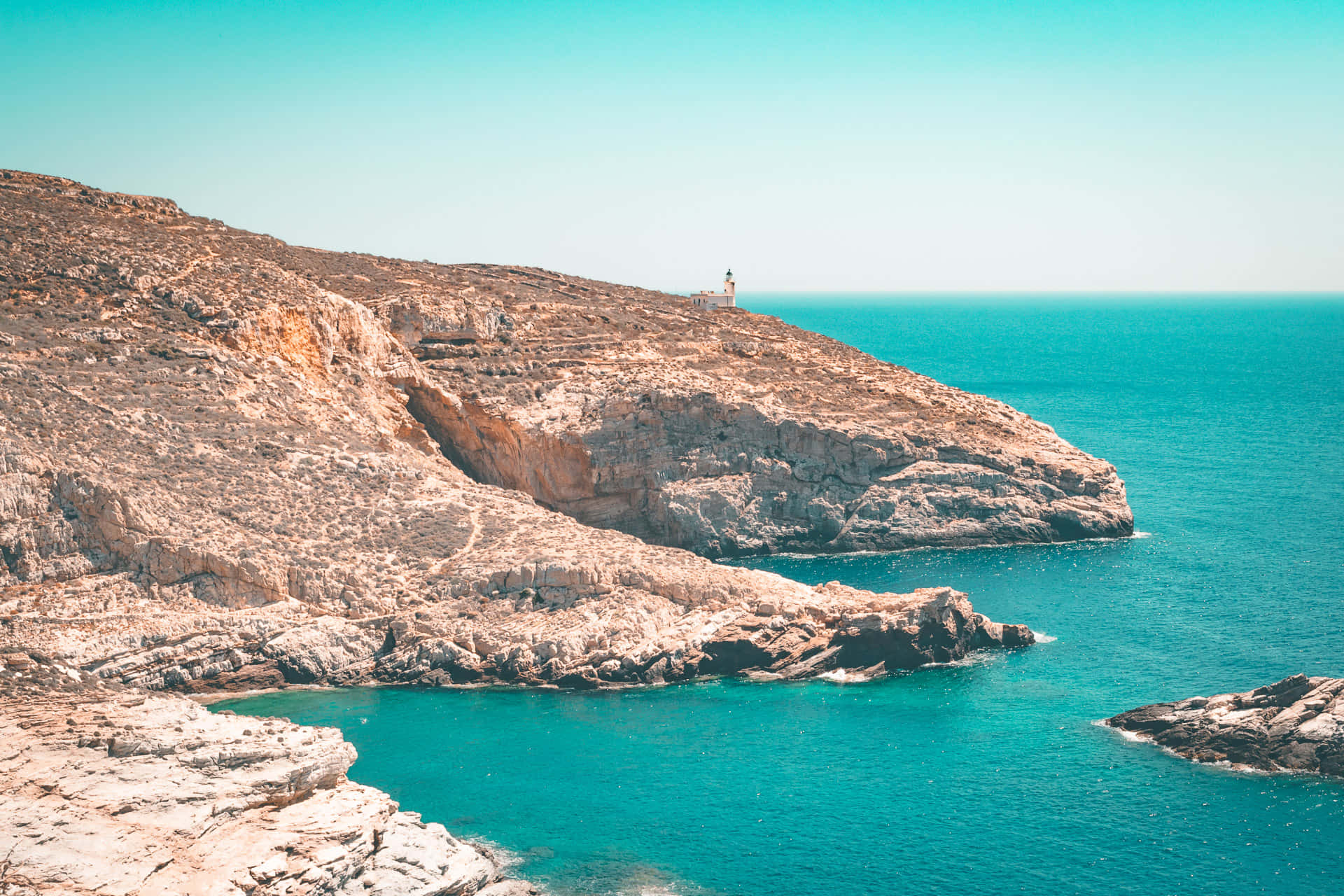 Greek Beach 桌布 壁紙 — 一座燈塔棲息在崎嶇的懸崖上，俯瞰著清澈藍天下充滿活力的碧綠海水。