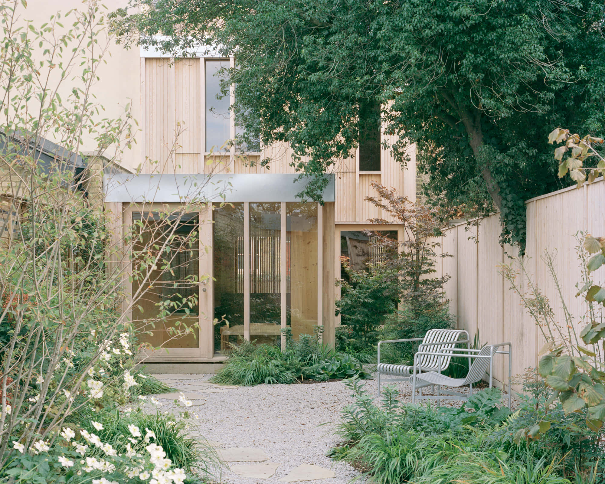 Minimalist Architecture 桌布 壁紙 — 一座現代木屋，配有玻璃牆，坐落於鬱鬱蔥蔥的綠色花園中，有碎石小徑和金屬椅子。