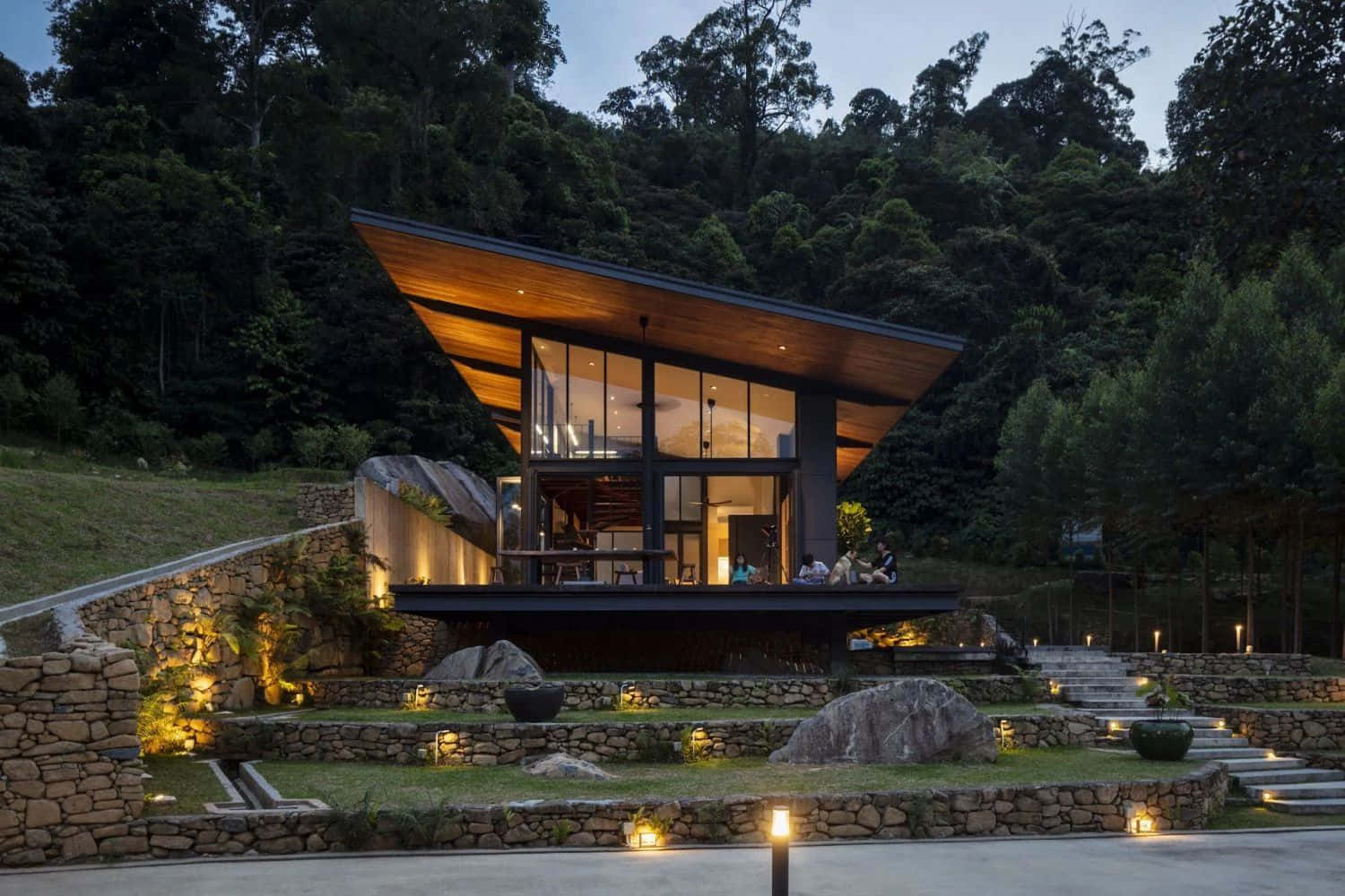 Minimalist Architecture 桌布 壁紙 — 一座現代木屋，配有玻璃牆和溫暖的室內燈光，坐落在黃昏時分的茂密森林中。