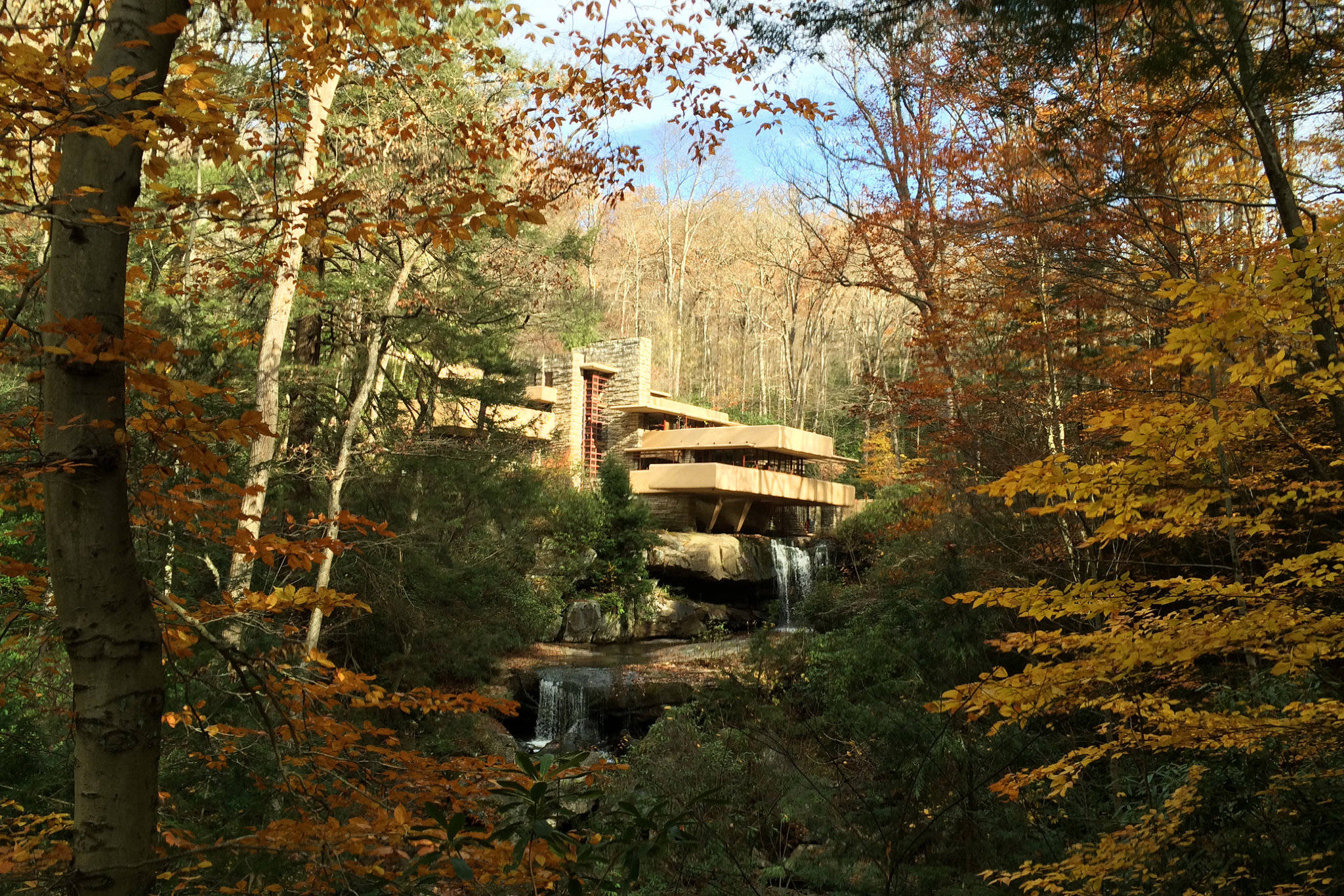 Falling Water 桌布 壁紙 — 一座現代木屋坐落在瀑布旁的岩石上，周圍是秋日樹木，金黃的樹葉映襯著清澈的藍天。