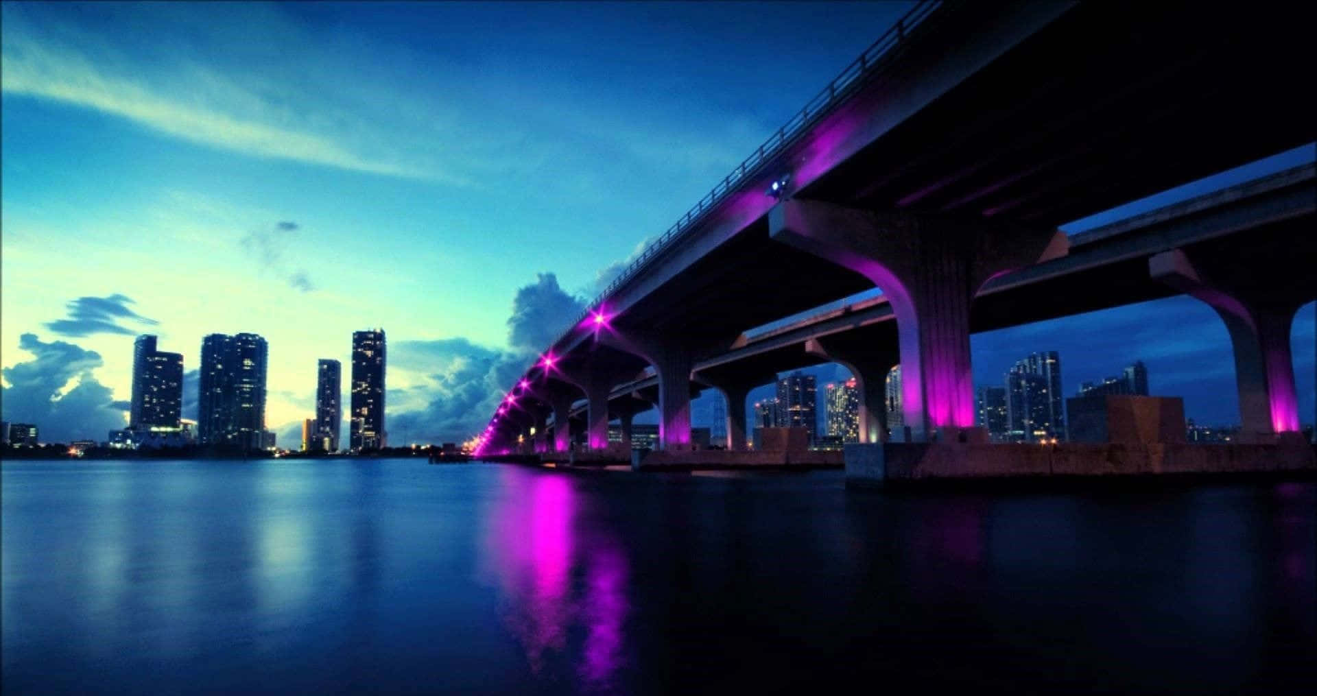 放鬆 桌布 壁紙 — 一座現代橋樑在暮色中橫跨平靜的水道，紫色燈光倒映在閃爍摩天大樓的城市天際線上。