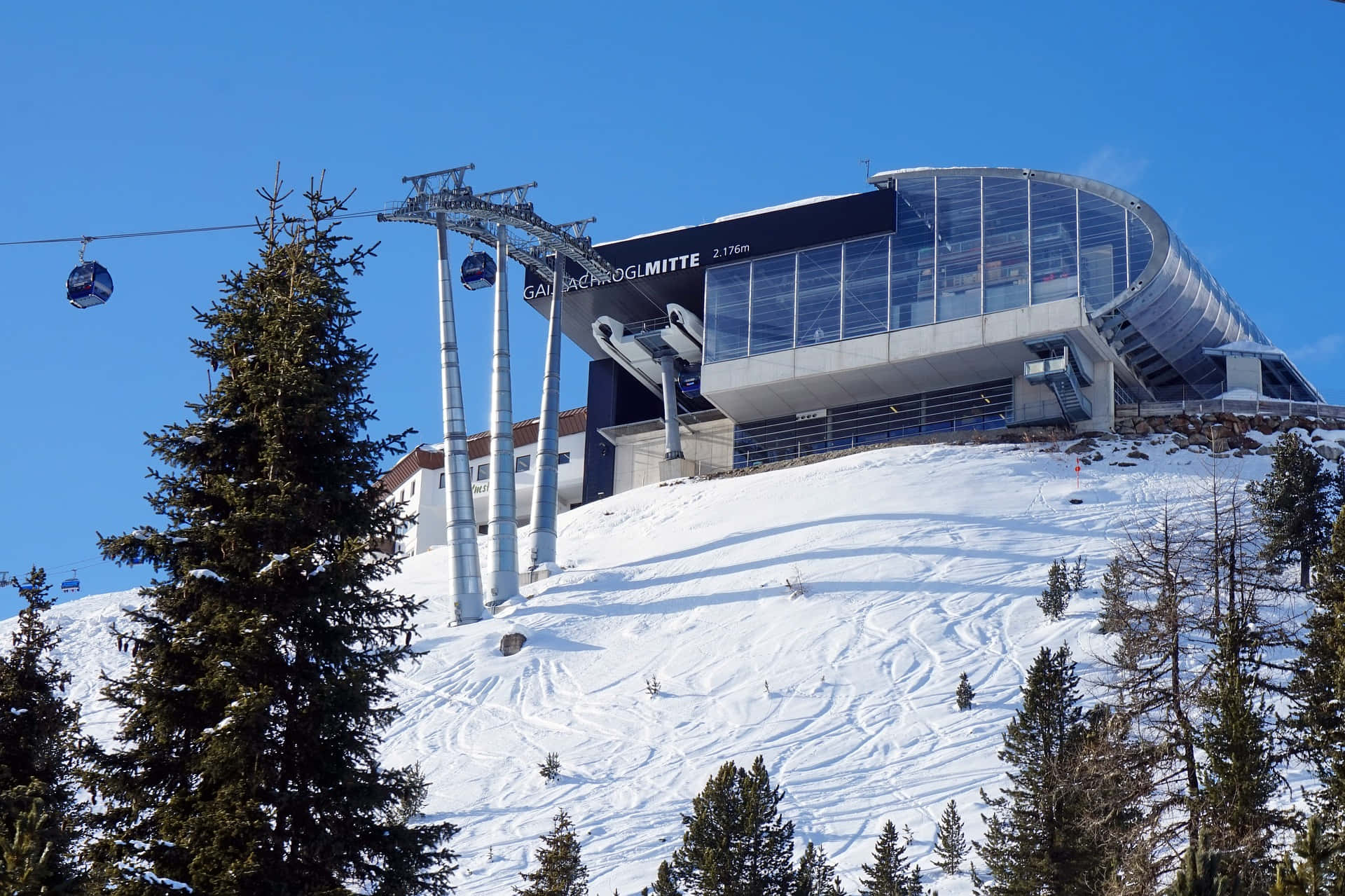 Ski Resort 桌布 壁紙 — 一座現代玻璃滑雪纜車站，坐落在白雪覆蓋的山頂上，藍天清澈，周圍有常青樹。