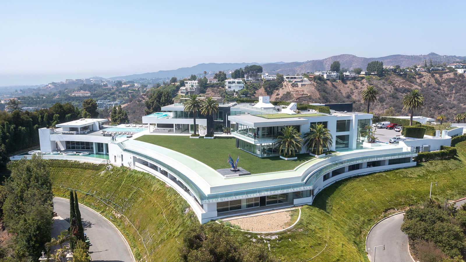 Big House 桌布 壁紙 — 一座現代白色豪宅的鳥瞰圖，擁有綠色屋頂和游泳池，坐落在綠草如茵的山坡上，俯瞰遠處的山丘。