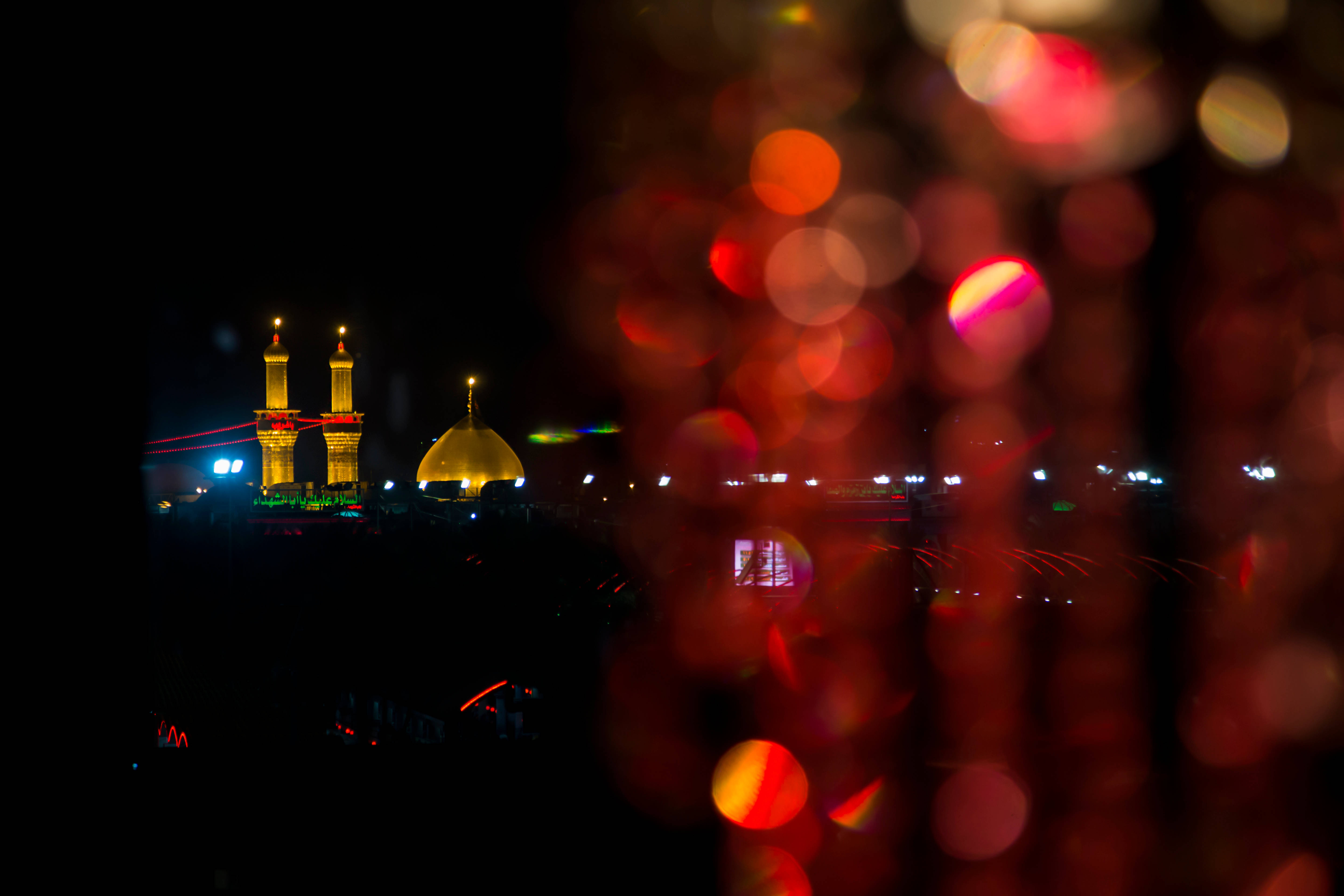 Karbala 桌布 壁紙 — 一座發光的清真寺，帶有金色圓頂和宣禮塔，在夜晚透過溫暖的紅色散景燈光前景閃耀。