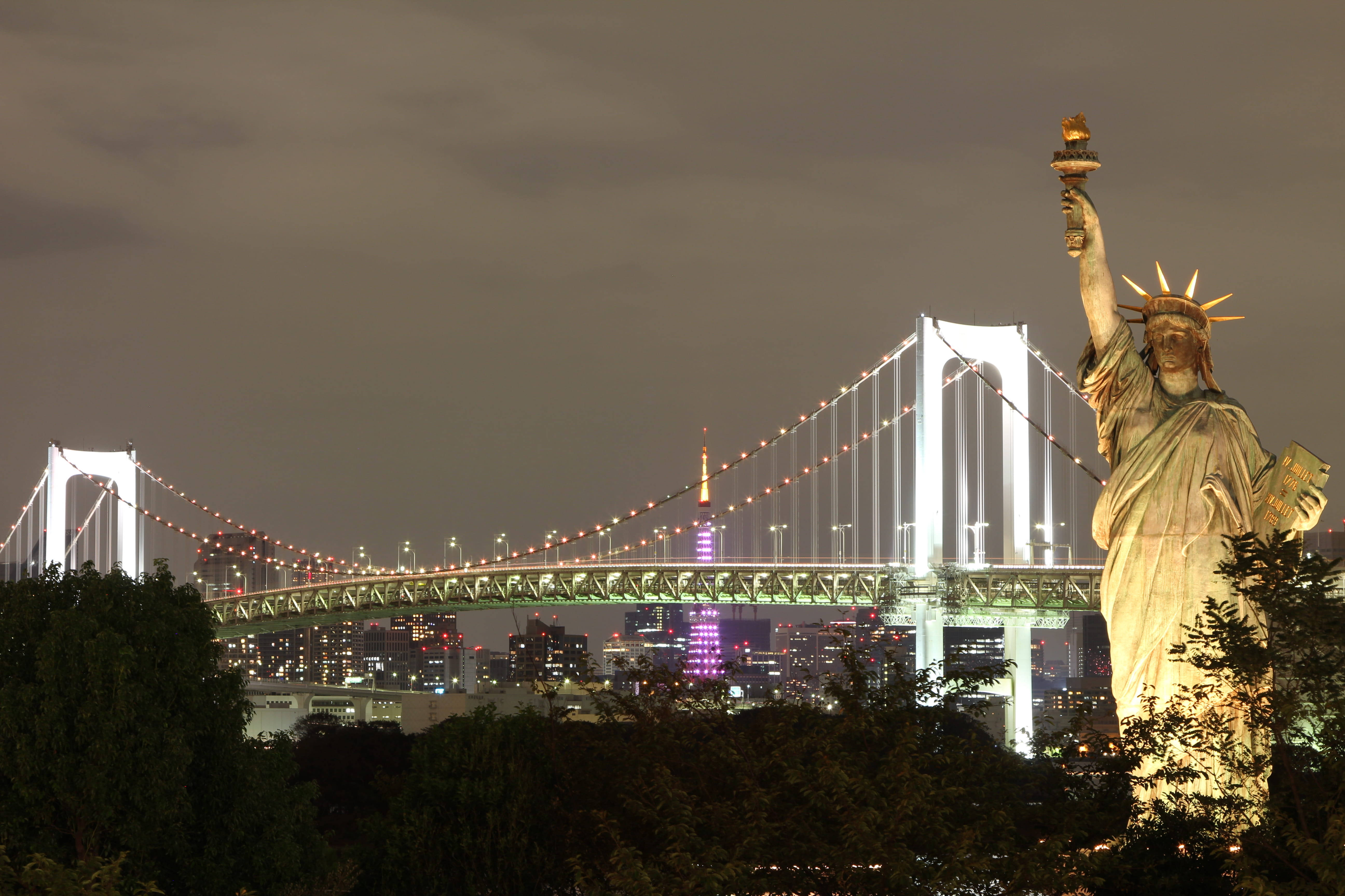 new york 4K 桌布 壁紙 — 一座發光的自由神像複製品在夜晚矗立於東京亮燈的彩虹橋前。