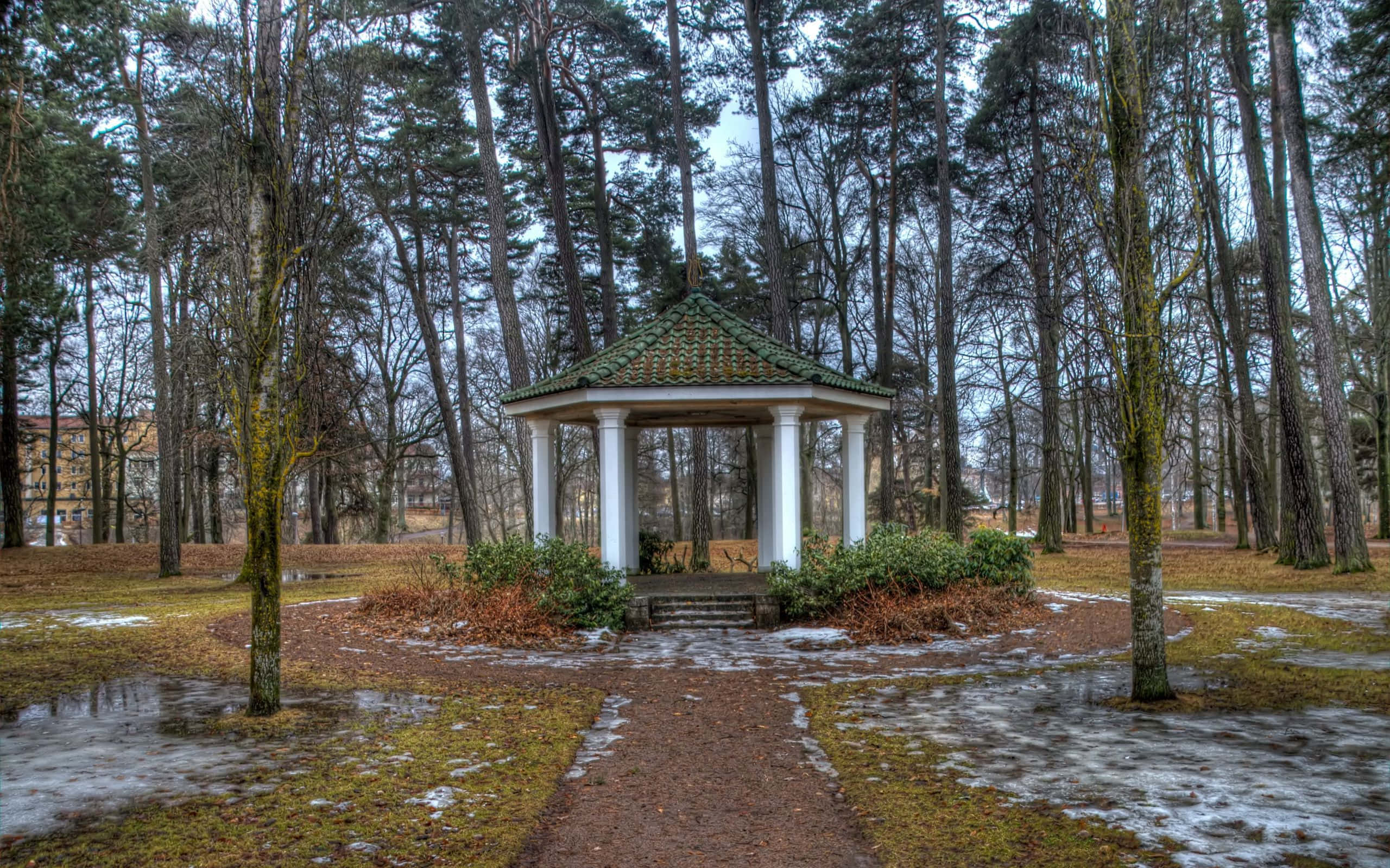 Spring Park 桌布 壁紙 — 一座白色柱式涼亭，配以綠色瓦片屋頂，矗立於白雪覆蓋的松樹林中，有一條泥濘小徑通往該處。