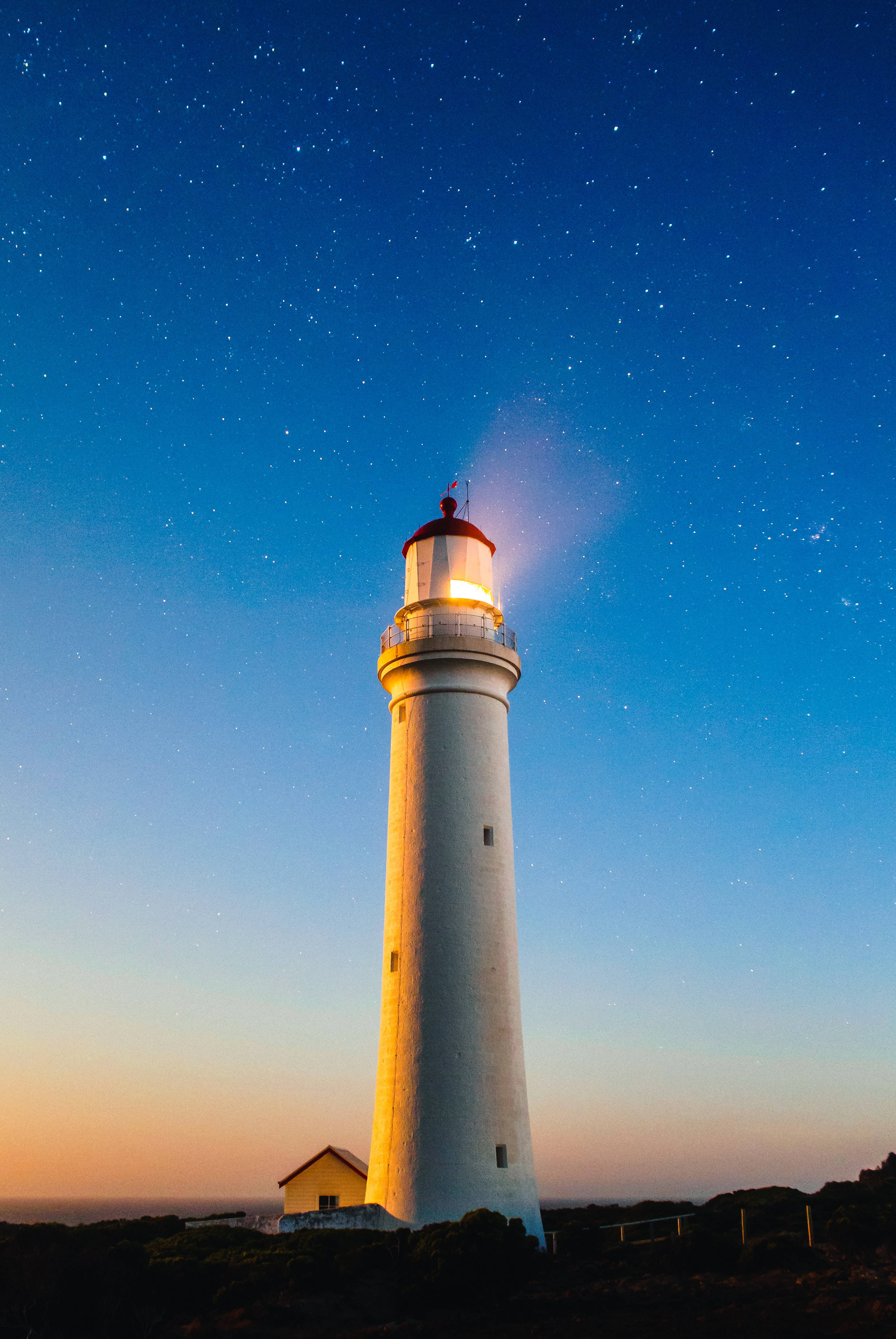 夜空 桌布 壁紙 — 一座白色燈塔在星空暮色中散發溫暖光芒，天空呈現藍色與橙色的漸變色調。