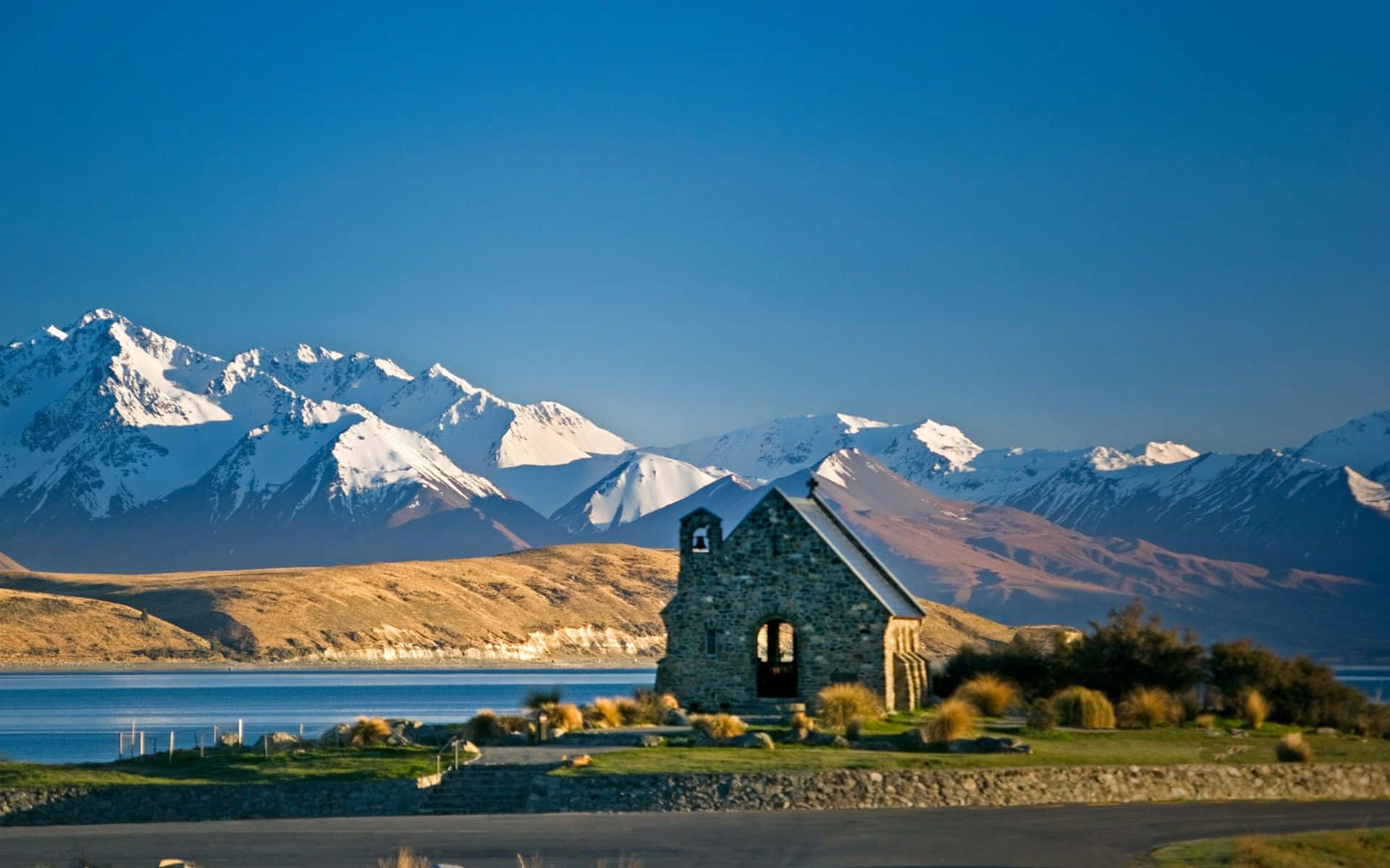 New Zealand 桌布 壁紙 — 一座石教堂獨自矗立在湖岸邊，背後是白雪覆蓋的山峰，在清澈的藍天下聳立。