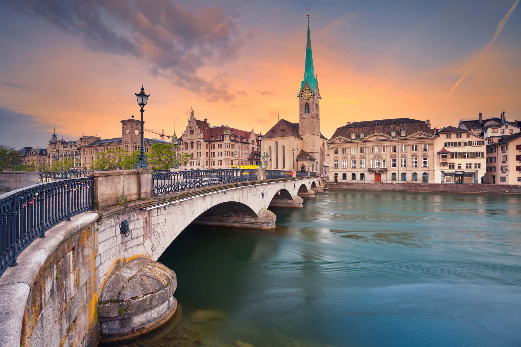 Switzerland 桌布 壁紙 — 一座石橋在日落時分拱跨平靜的河流，歷史建築和綠色尖塔在溫暖的光線中閃耀。