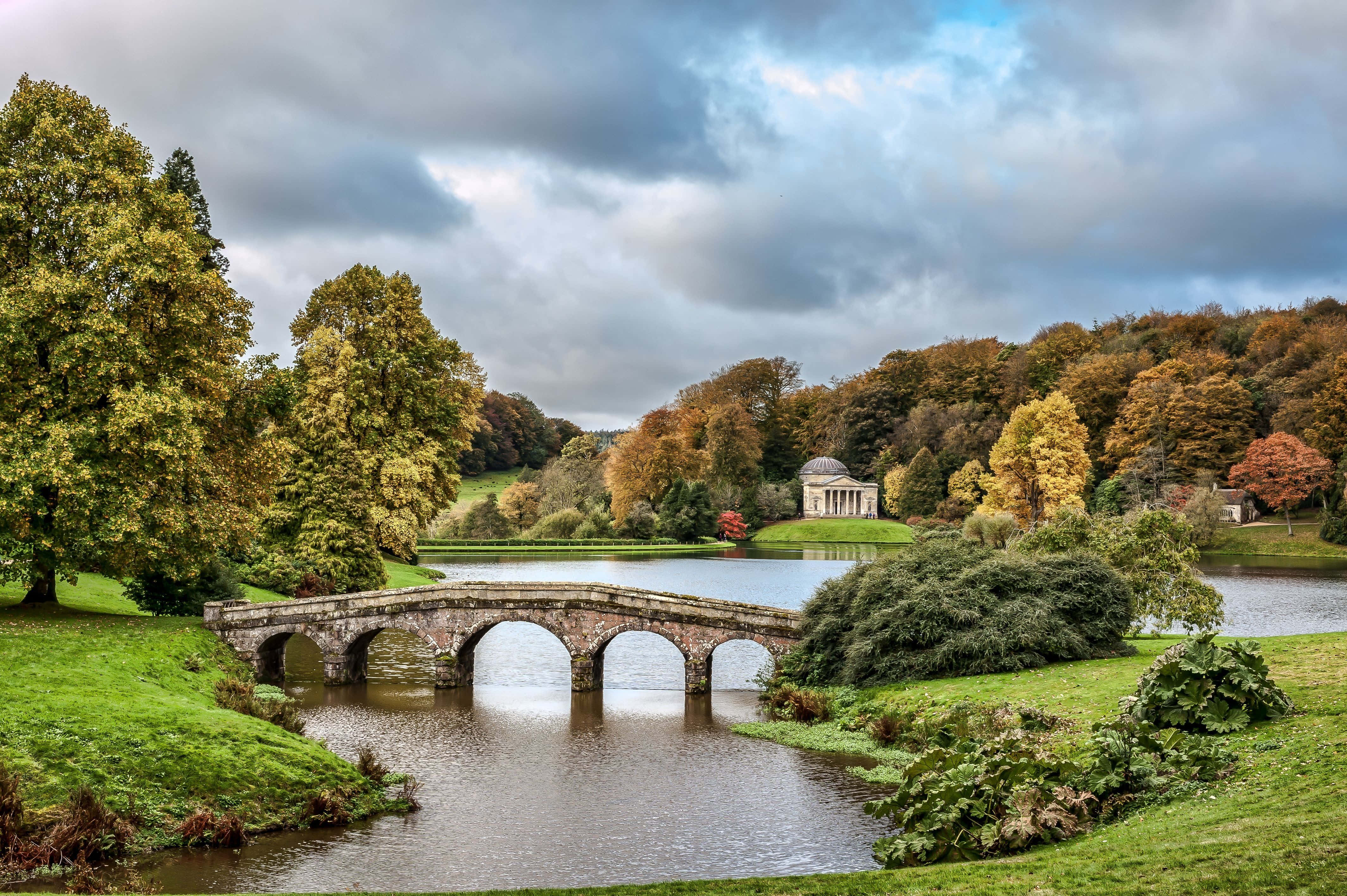UK 桌布 壁紙 — 一座石橋在秋日公園中拱跨平靜的河流，周圍有色彩繽紛的樹木，遠山上有古典神廟，天空多雲。