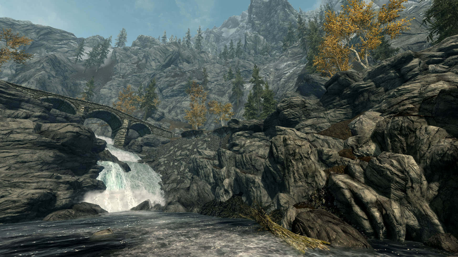 Skyrim Landscapes 桌布 壁紙 — 一座石橋拱起於奔騰的瀑布之上，坐落於崎嶇的山谷中，周圍有秋天的樹木。