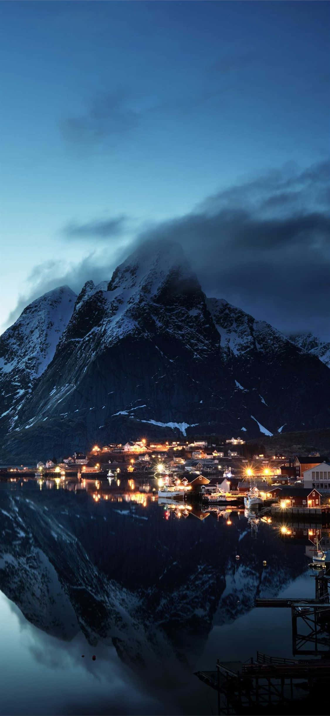 Norway 桌布 壁紙 — 一座積雪的山峰聳立於峽灣小鎮之上，小鎮溫暖的燈光在暮色天空下倒映在平靜的水面上。
