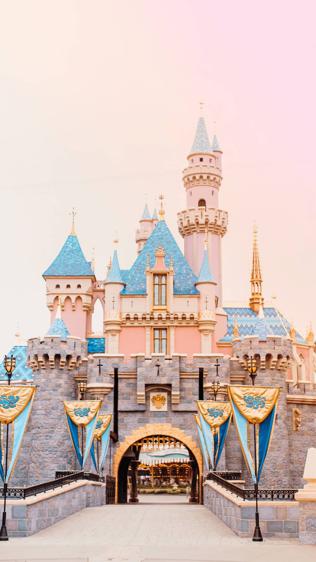 Disneyland Castle 桌布 壁紙 — 一座童話城堡，粉紅色的牆壁和藍色的屋頂，矗立在柔和的粉紅色天空下，入口處懸掛著旗幟。