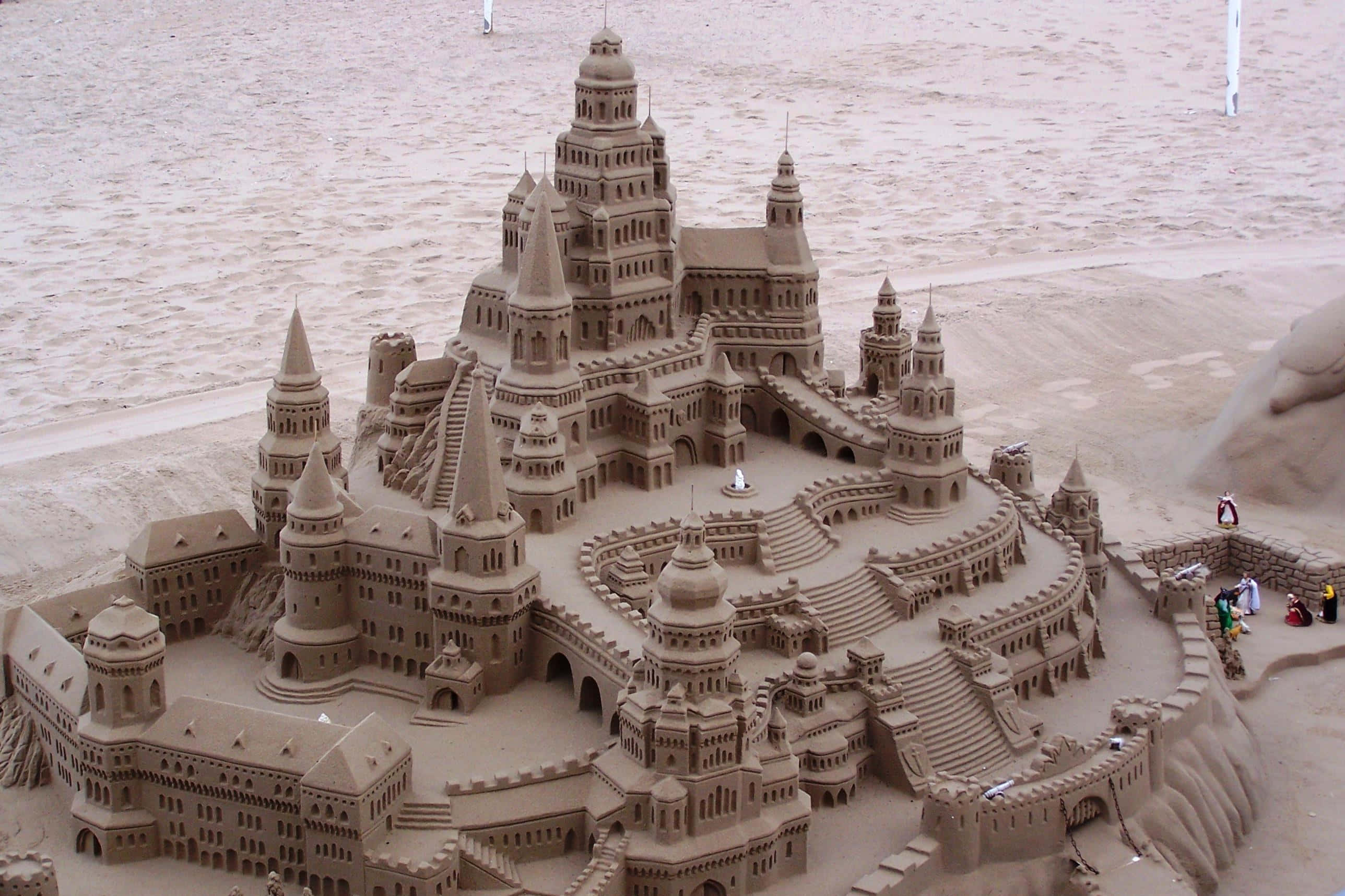 Sandcastles 桌布 壁紙 — 一座精緻的沙堡要塞，帶有塔樓和橋樑，建在沙灘上，附近有人。