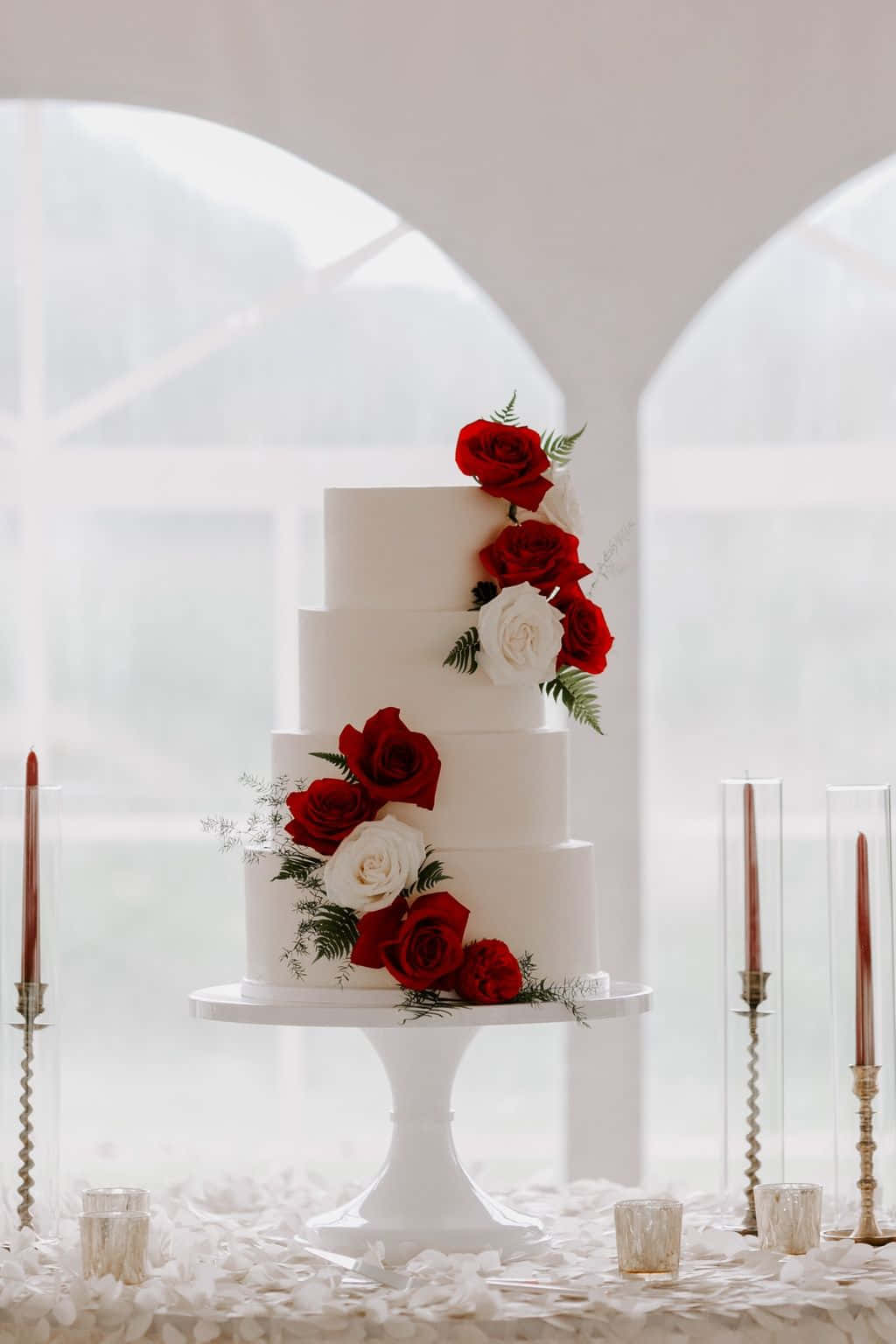Roses Wedding 桌布 壁紙 — 一座純白多層婚禮蛋糕，飾以紅白玫瑰瀑布，優雅地立於拱形窗旁的基座上。