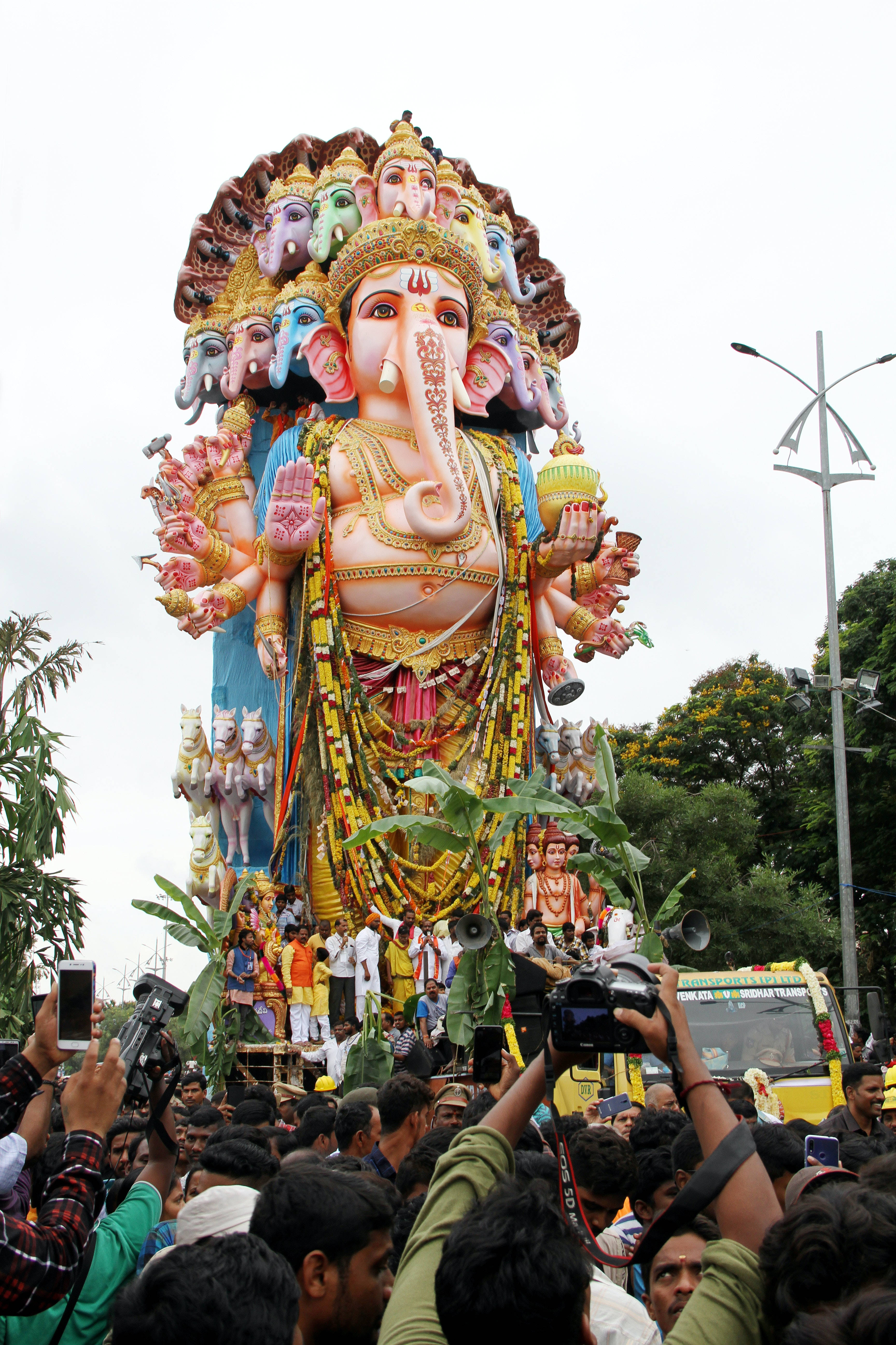 Ganesh Chaturthi 桌布 壁紙 — 一座色彩繽紛、多頭多臂的象神像在擠滿人的街道上巡遊，信徒和攝影師在下方聚集。