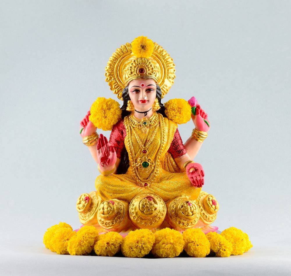 Lakshmi 桌布 壁紙 — 一座色彩鮮豔的多臂印度教女神金色雕像，飾有萬壽菊和珠寶，寧靜地坐在素色背景前。