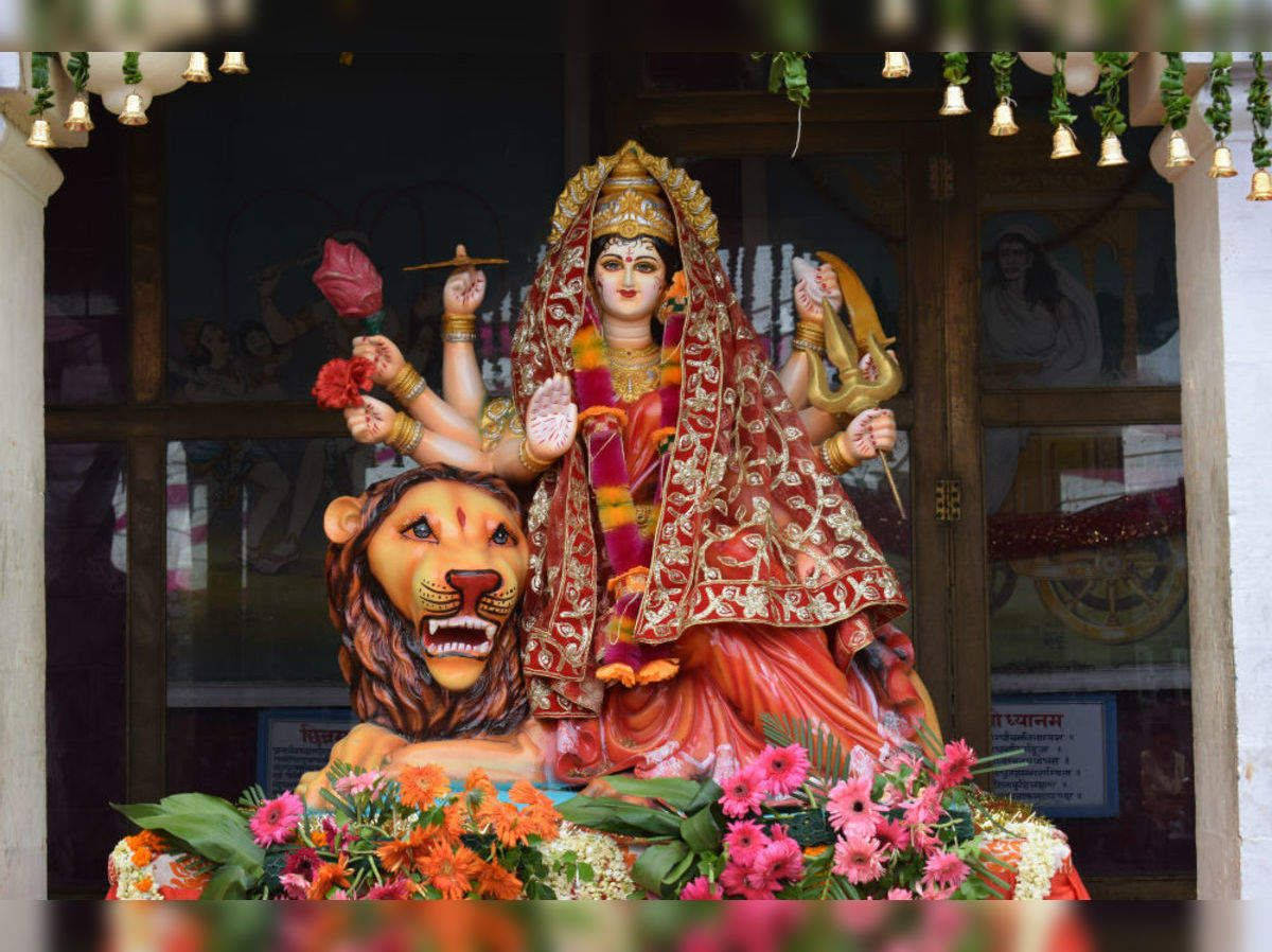 statue 桌布 壁紙 — 一座色彩鮮豔的多臂女神雕像，身著紅金色，坐在一頭咆哮的獅子上，周圍環繞著色彩繽紛的花朵和懸掛的鈴鐺。