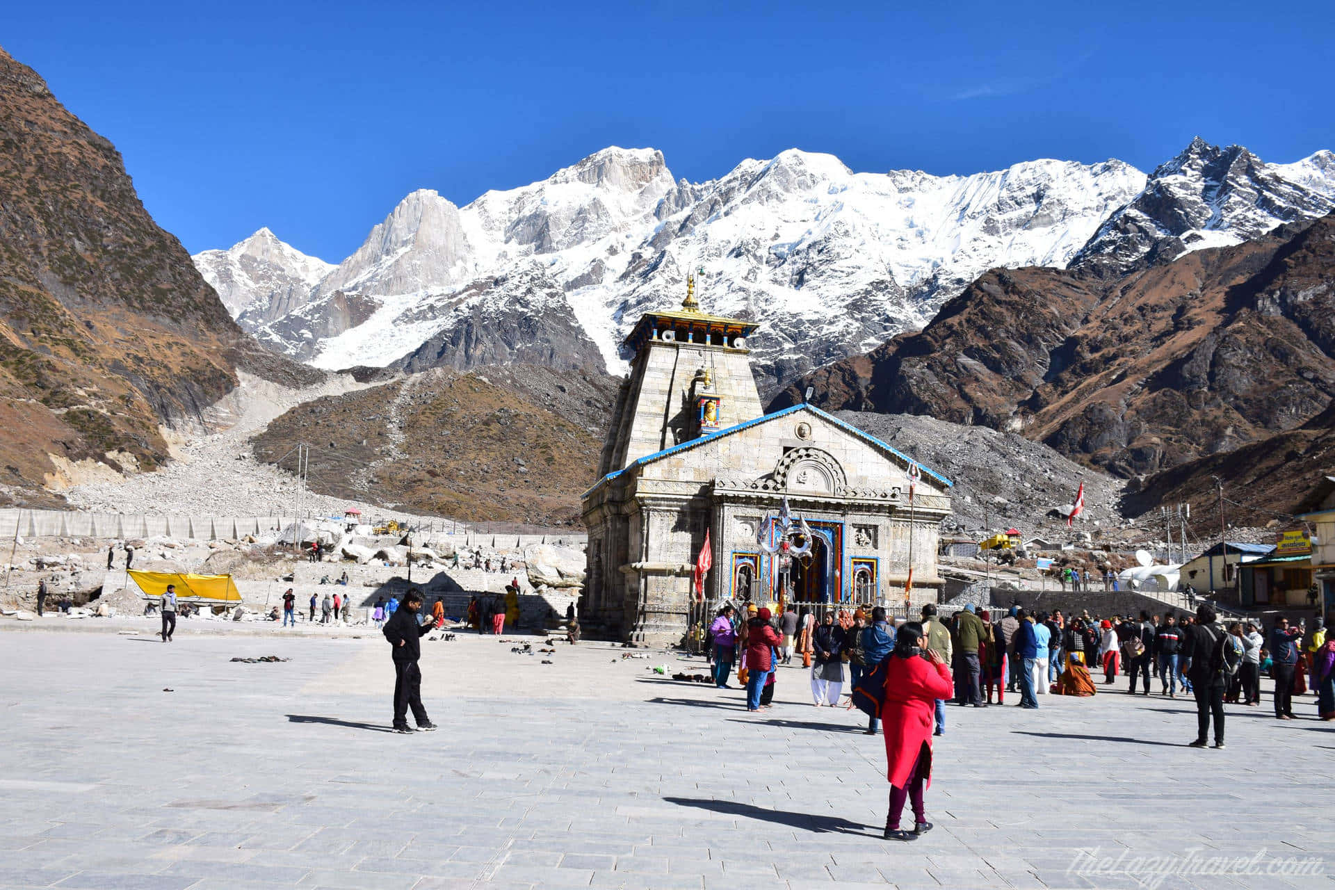 Kedarnath 桌布 壁紙 — 一座色彩鮮豔的寺廟矗立在白雪覆蓋的高聳山脈前，藍天白雲下，朝聖者們走過石頭廣場。