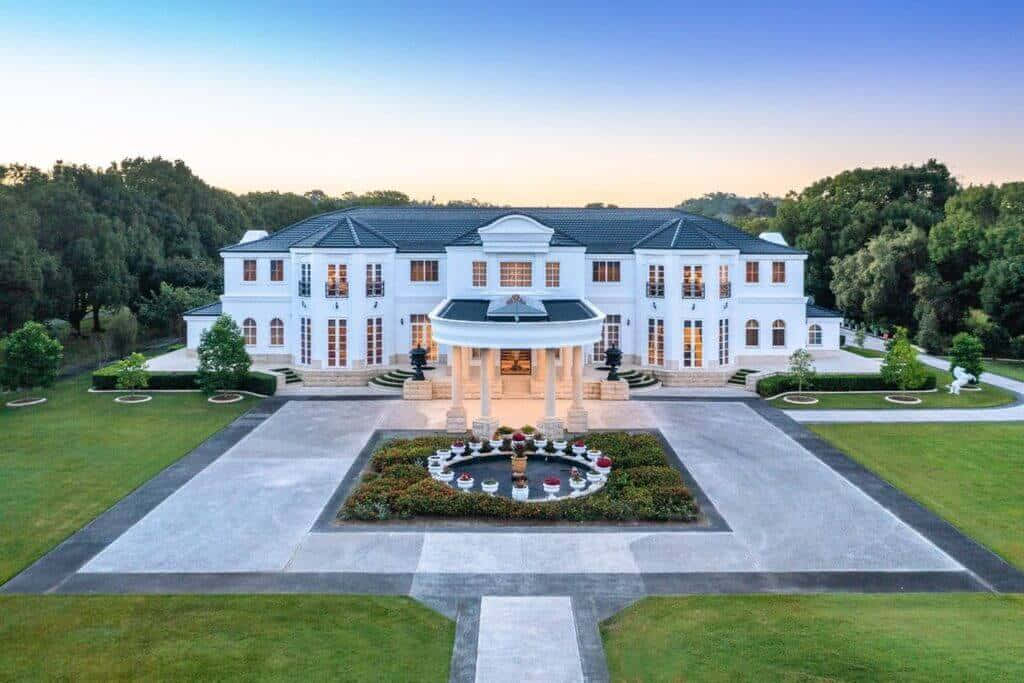 一座被繁茂綠意環抱的美麗房屋 壁紙 — 一座宏偉的白色大宅，中央有噴泉和對稱花園，沐浴在金色的日落光芒中。