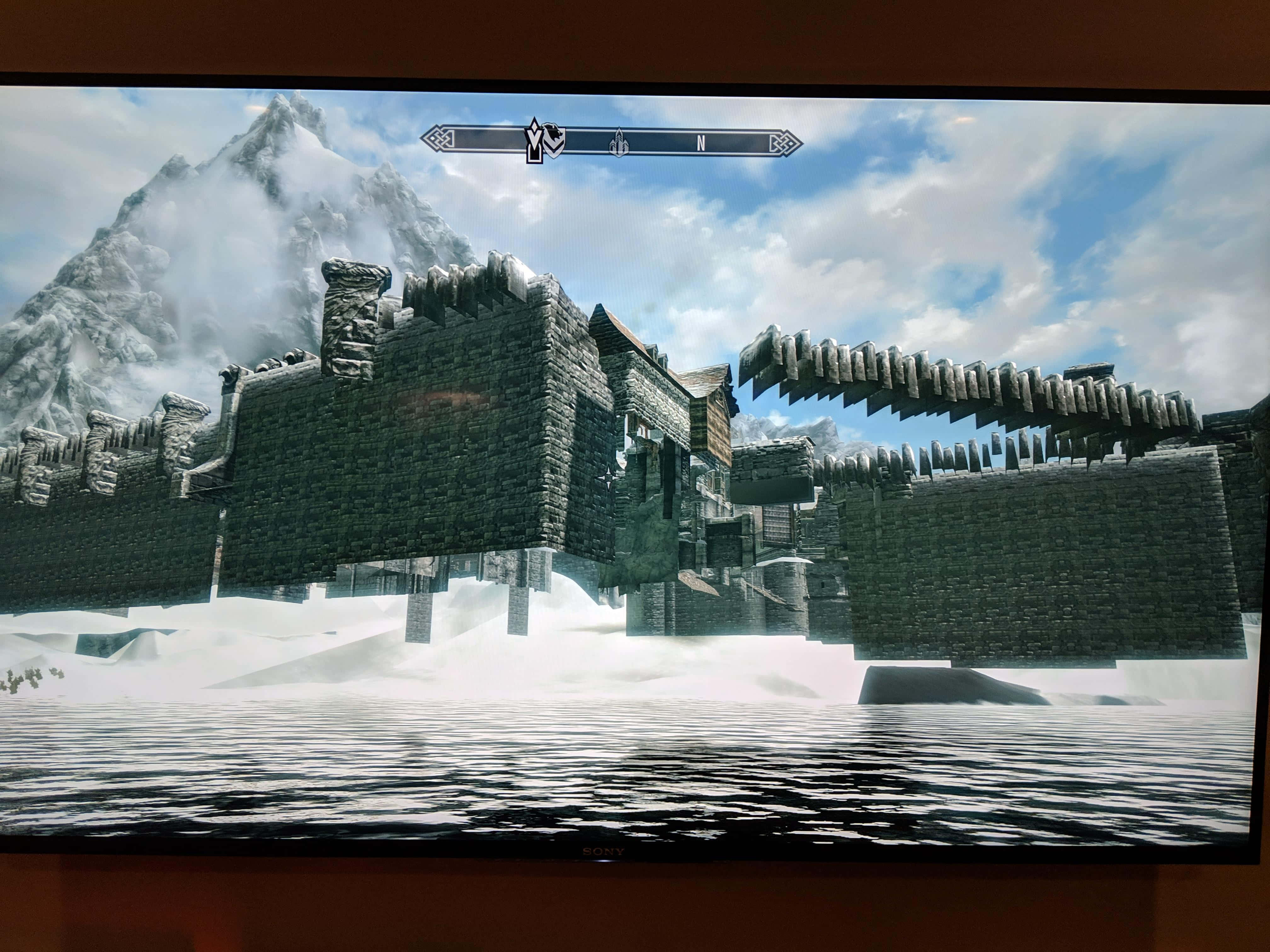 Skyrim Windhelm 桌布 壁紙 — 一座被雪覆蓋的山上堡壘，擁有石牆和冰橋，漂浮在結冰的湖面上，天空多雲。