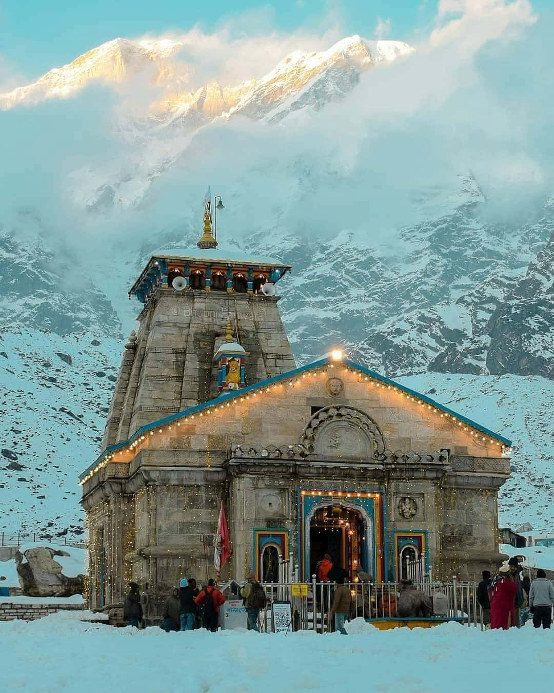 Kedarnath 桌布 壁紙 — 一座被雪覆蓋的石廟在柔和的藍天下，對著雄偉的山脈溫暖地發光。