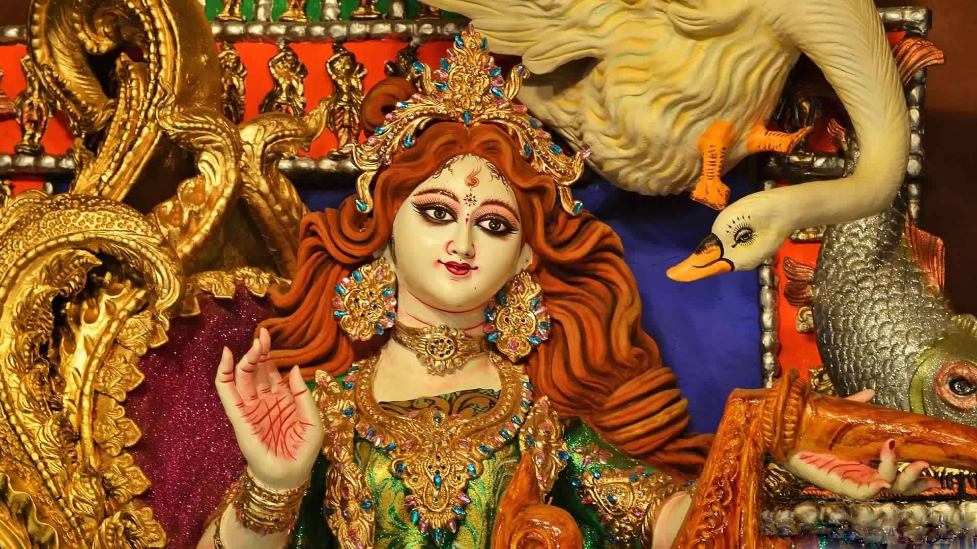Saraswati 桌布 壁紙 — 一座裝飾華麗的印度教女神雕像，擁有飄逸的紅髮和金色珠寶，坐在一隻白天鵝旁邊，背景是鮮豔的紅色和藍色。