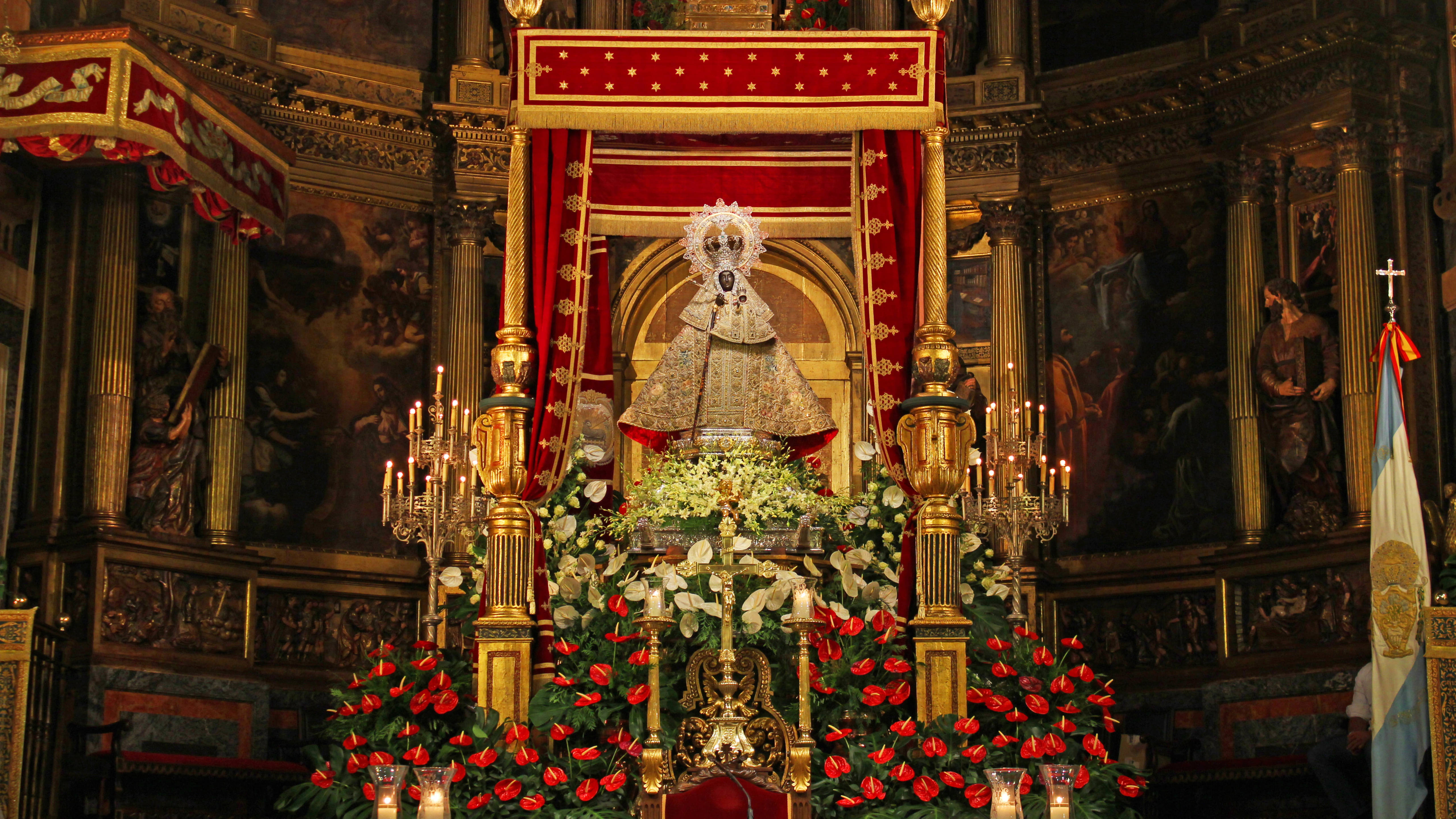 Virgen De Guadalupe 桌布 壁紙 — 一座裝飾華麗的宗教雕像端坐在紅色華蓋下，周圍環繞著金色燭台和鮮豔的紅白花朵，位於宏偉華麗的小教堂內。