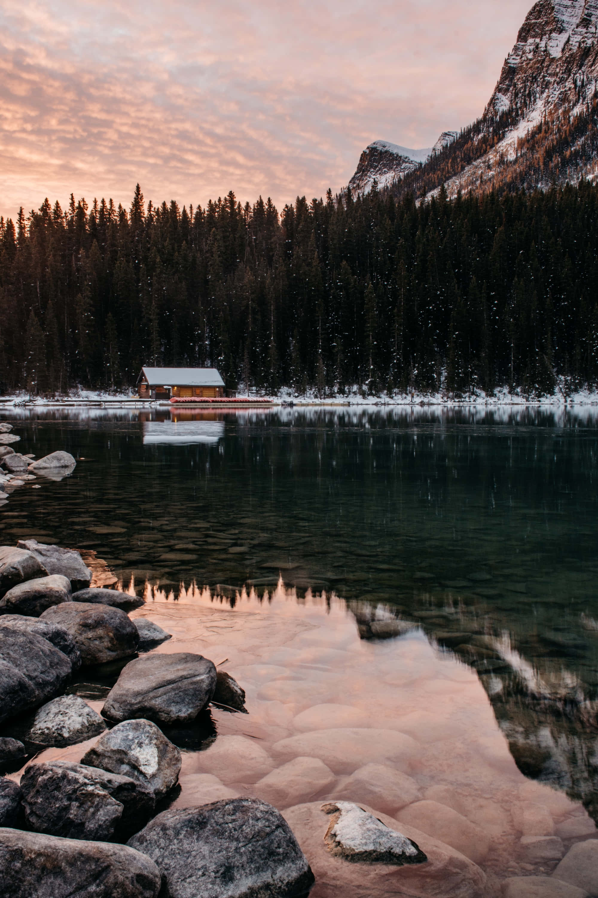 Phone Screen 桌布 壁紙 — 一座覆蓋著薄雪的 cabin 在如鏡的湖面上，倒映出被常青森林和岩石海岸環繞的熾熱日落天空。