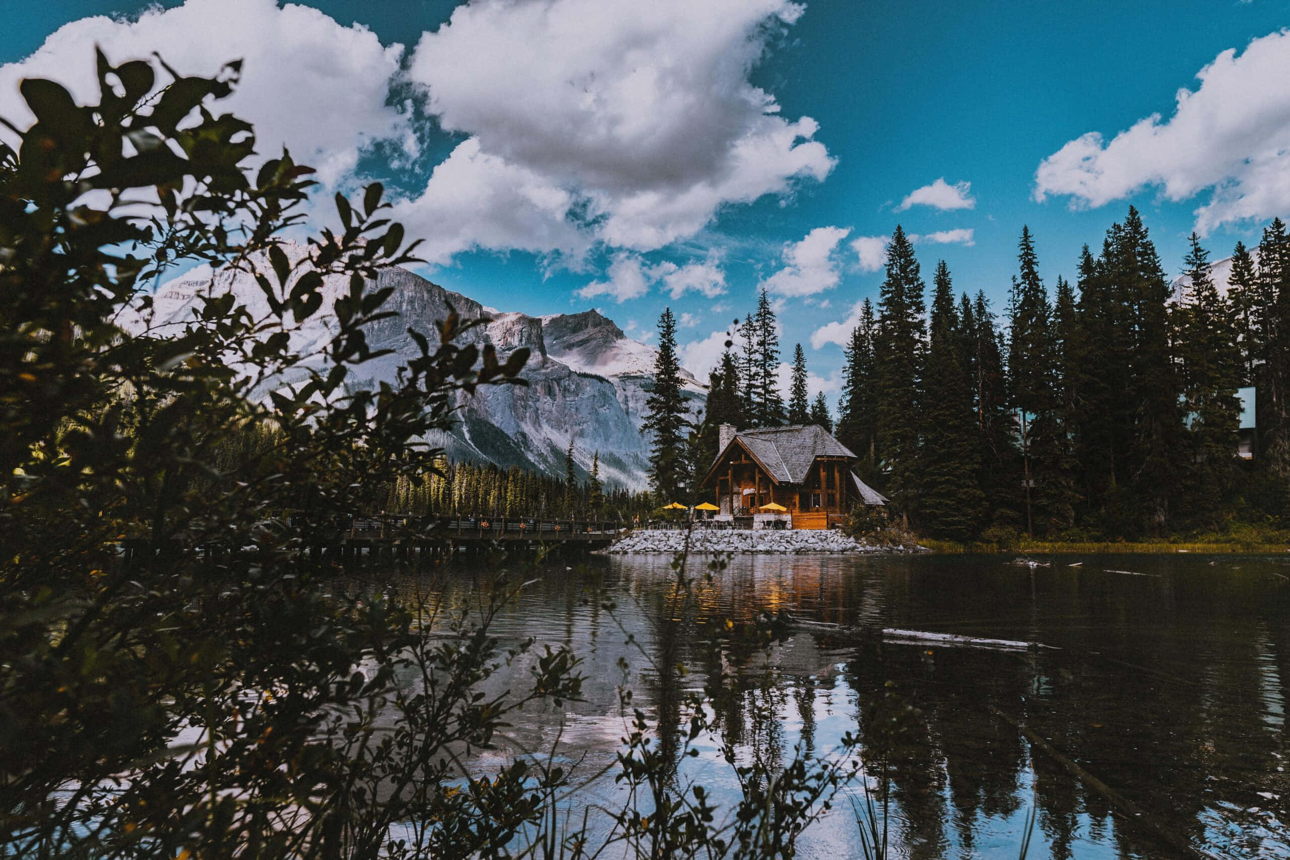 Nature Scenery 桌布 壁紙 — 一座質樸的木屋靜靜地坐落在湖岸邊，白雪覆蓋的山脈和松樹倒映在平靜的水面上，天空湛藍，白雲朵朵。