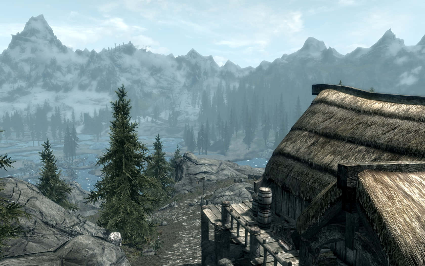 Skyrim Solitude 桌布 壁紙 — 一座質樸的茅草屋頂小屋坐落在岩石山坡上，俯瞰著霧氣瀰漫的山谷，山谷中有積雪覆蓋的山峰和常綠樹木。
