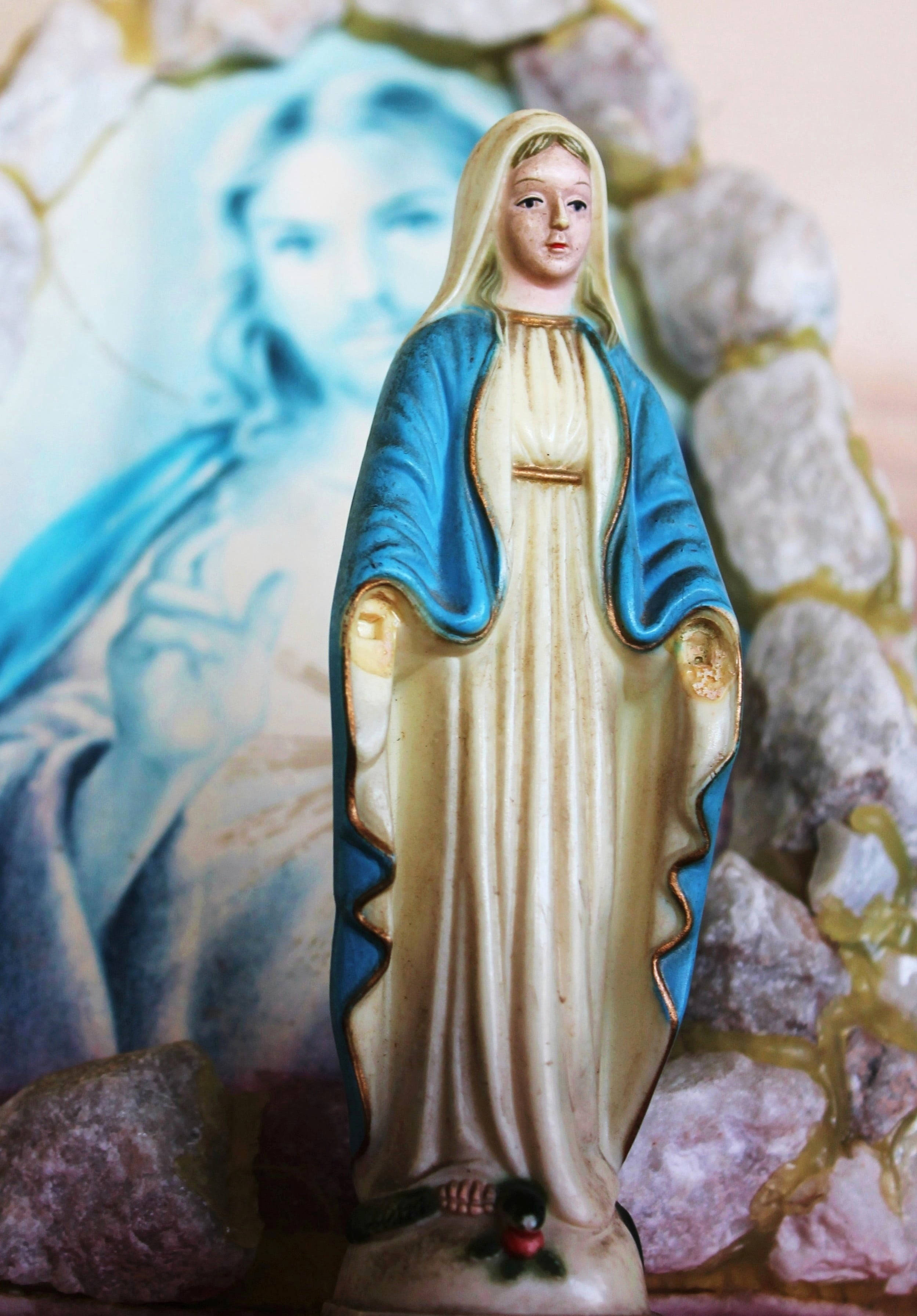 Virgen De Guadalupe 桌布 壁紙 — 一座身穿藍色和金色長袍的聖母瑪利亞寧靜雕像，矗立在模糊的宗教壁畫和石頭背景前。