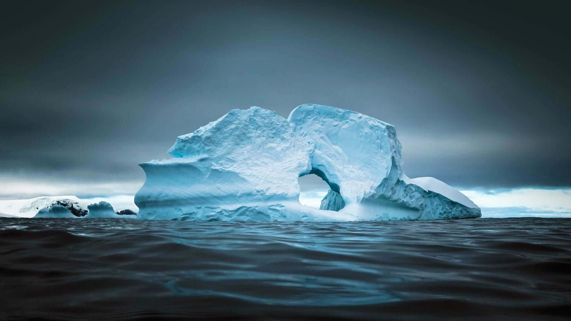 Antarctica 桌布 壁紙 — 一座雄偉的冰山帶有天然拱門，在陰沉的灰色天空下，靜靜地漂浮在黑暗的水面上。