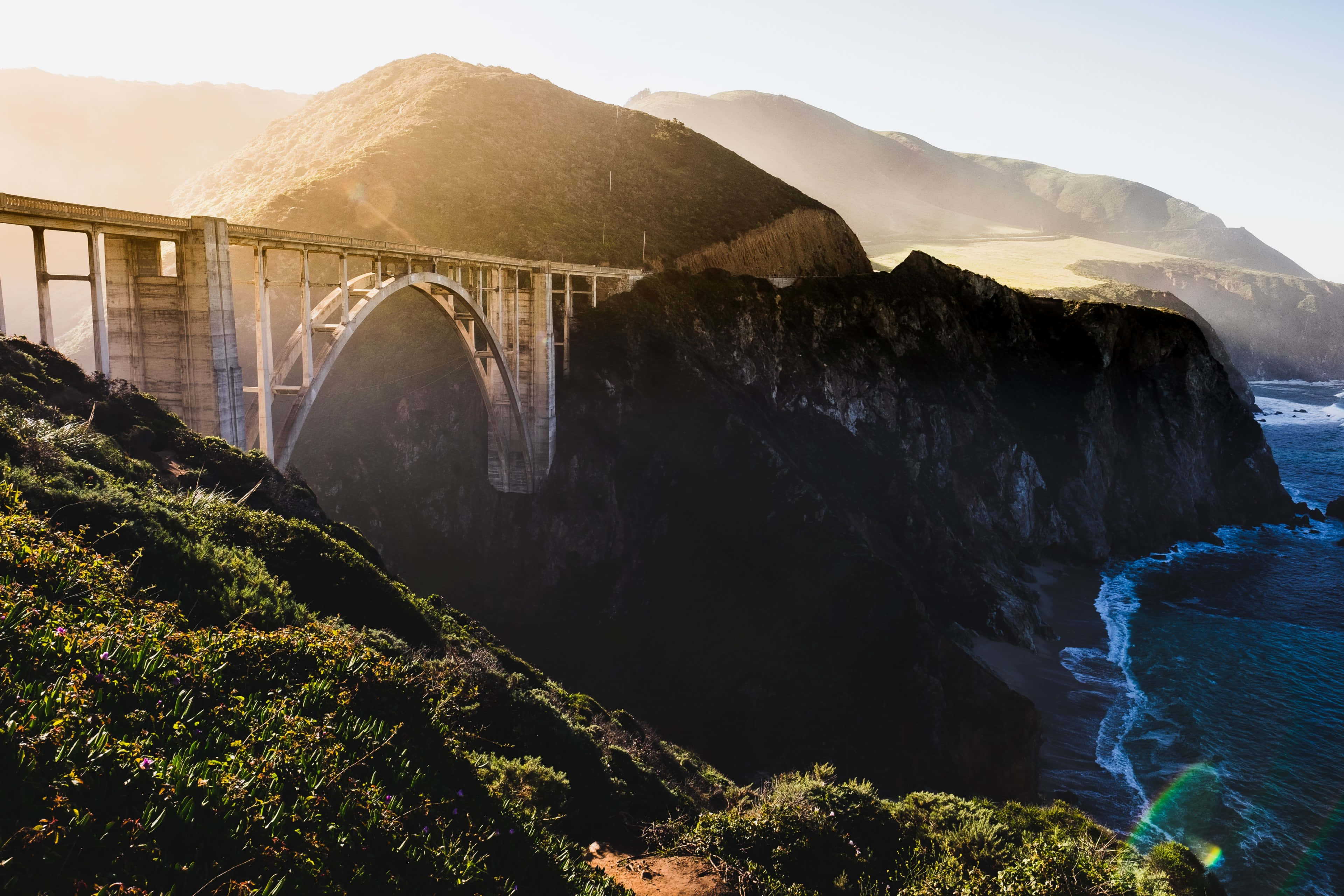 Big Sur 桌布 壁紙 — 一座雄偉的混凝土拱橋橫跨崎嶇的懸崖，俯瞰金色時刻翻騰的海浪。