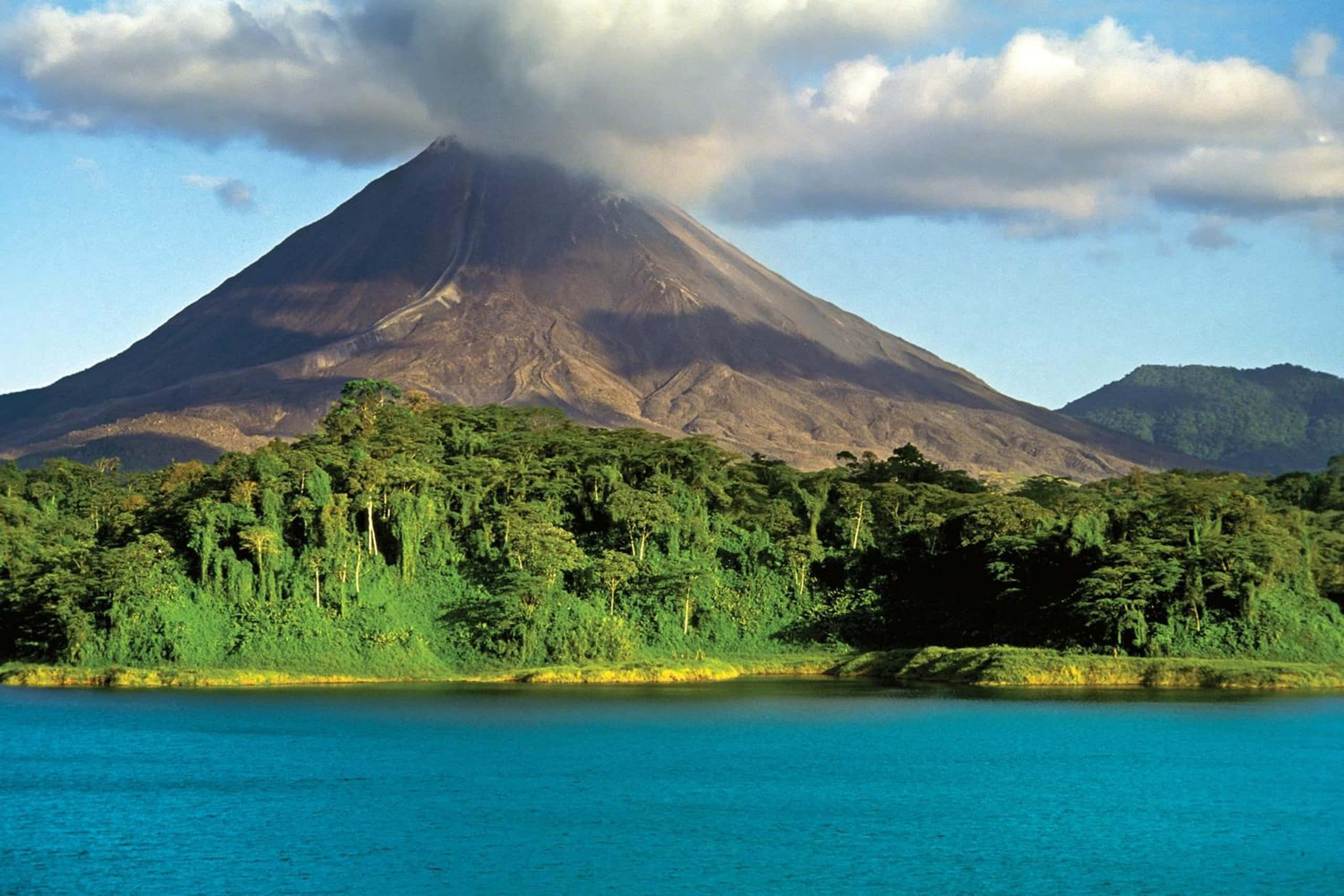 Costa Rica 桌布 壁紙 — 一座雄偉的火山山脈聳立在鬱鬱蔥蔥的綠色森林之上，森林毗鄰平靜的綠松石色湖泊，天空部分多雲。