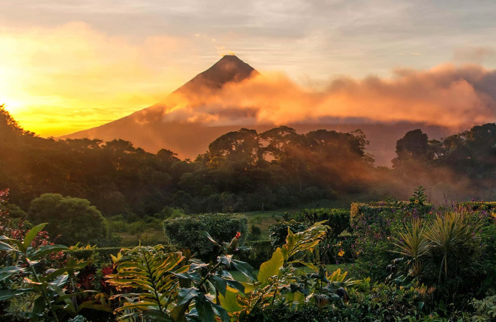 Costa Rica 桌布 壁紙 — 一座雄偉的火山穿過霧氣雲層升起，金色日出光芒沐浴著下方茂密的熱帶叢林。