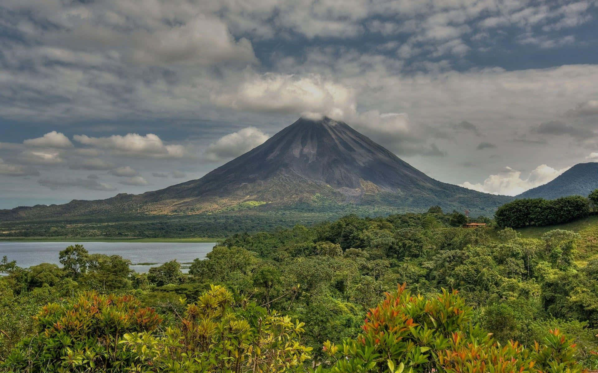 Costa Rica 桌布 壁紙 — 一座雄偉的火山聳立在茂密的綠色森林和寧靜的湖泊之上，天空多雲。