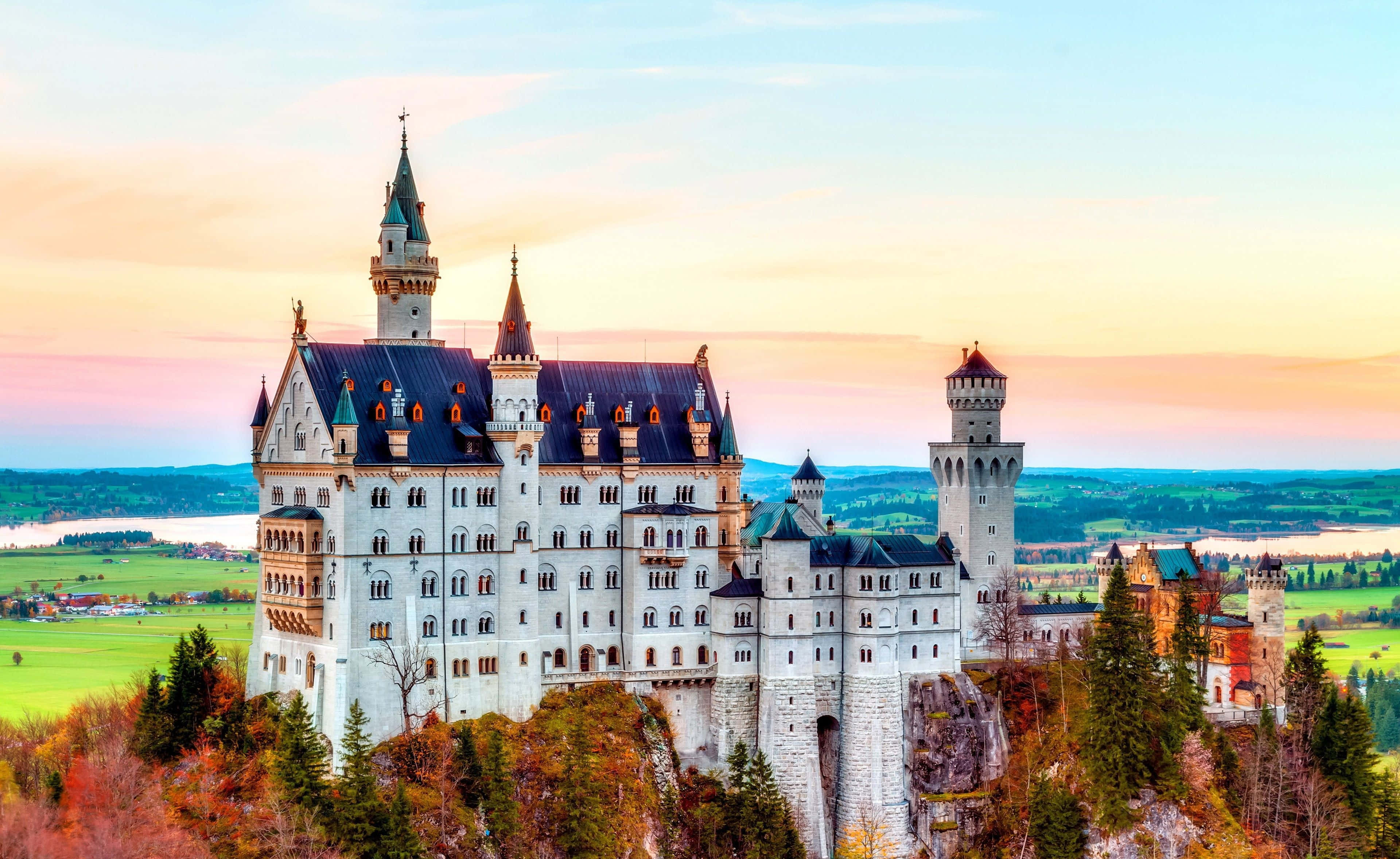 Germany 桌布 壁紙 — 一座雄偉的白色城堡，深色屋頂，矗立在岩石山丘上，周圍環繞著秋樹，在柔和的日落天空下。