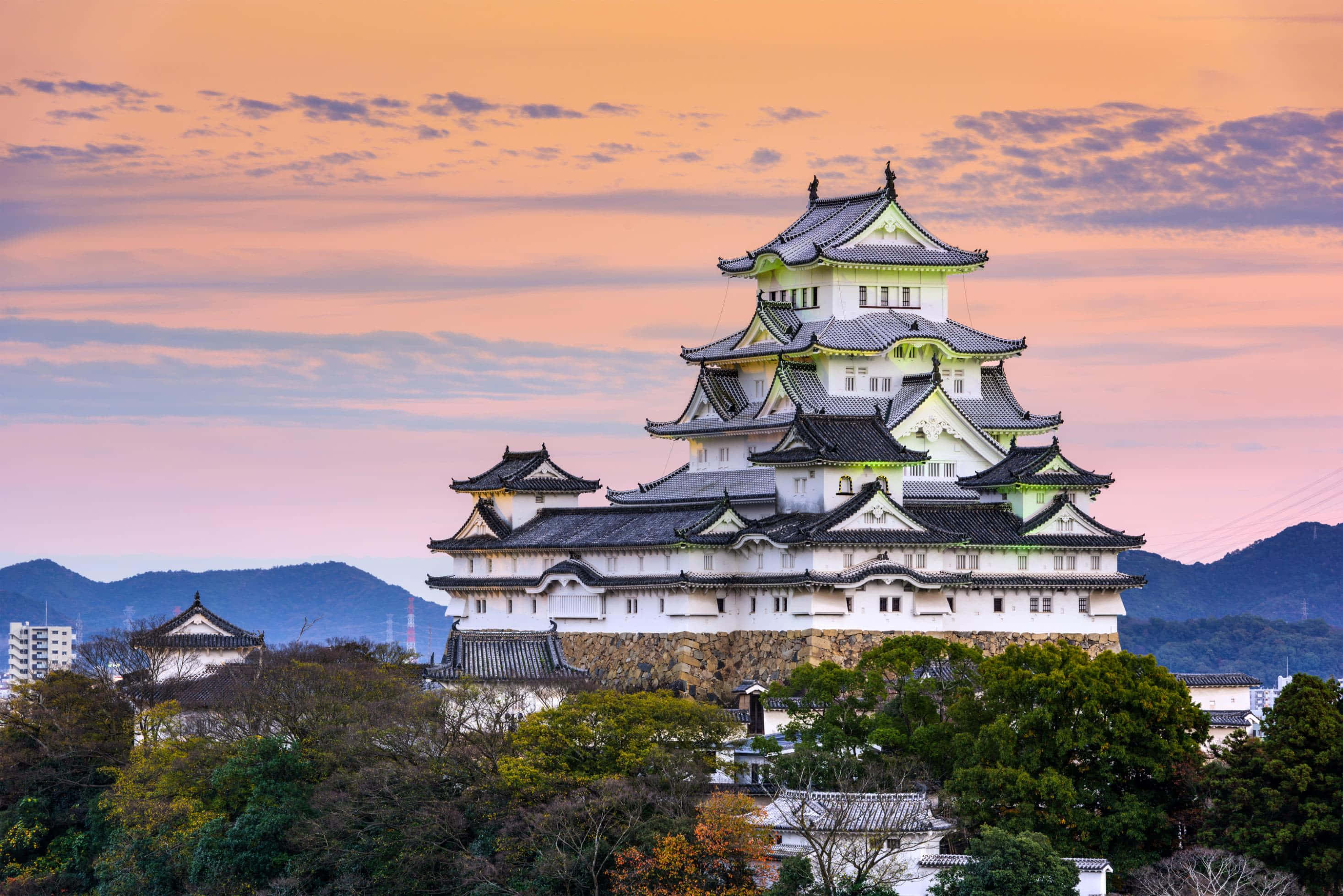 Japanese 桌布 壁紙 — 一座雄偉的白色日本城堡，帶有黑色瓦片屋頂，矗立在山丘上，夕陽西下，周圍環繞著茂密的綠樹和遠山，在溫暖的橙色天空下。
