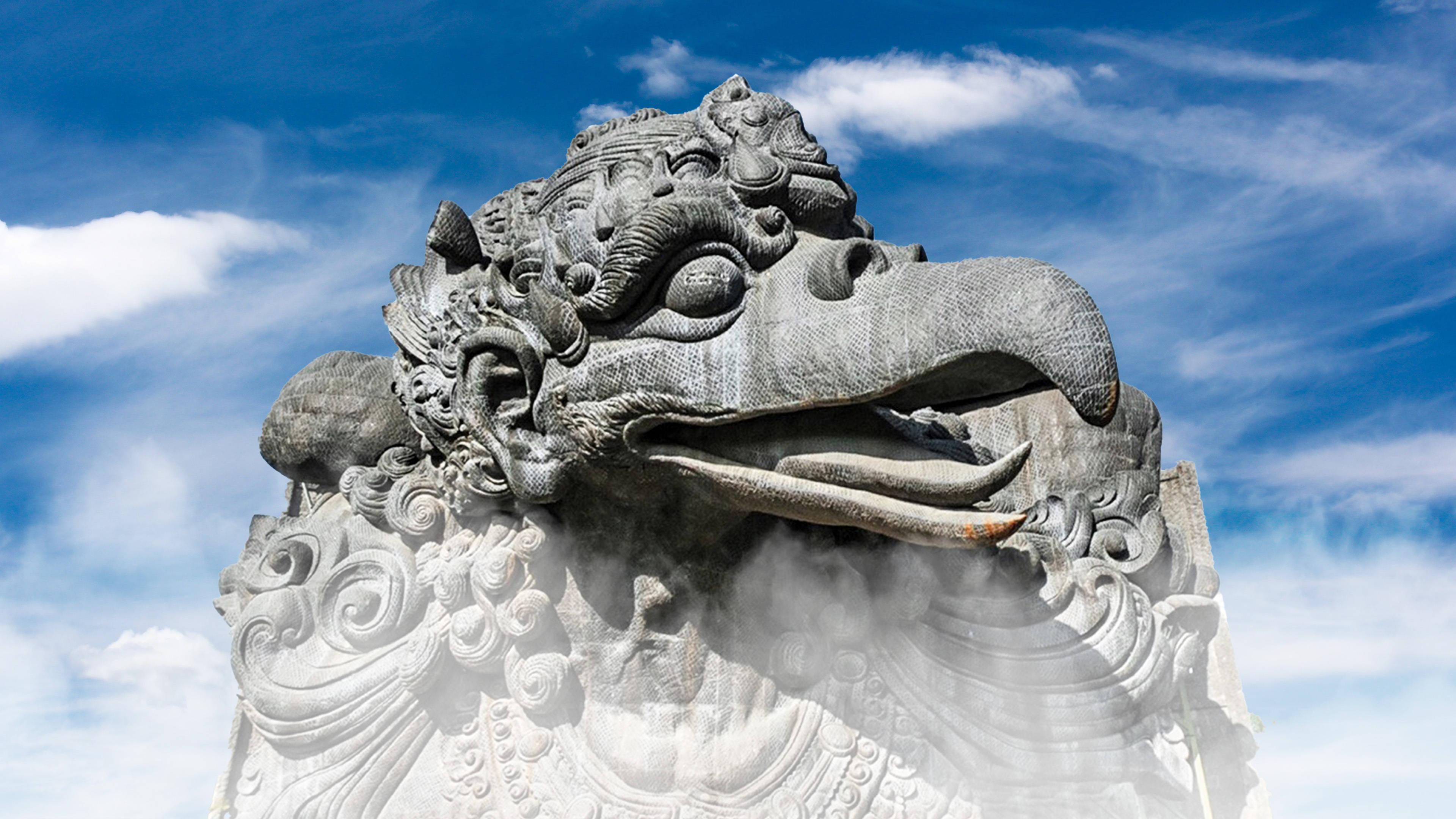 Statue Aesthetic 桌布 壁紙 — 一座雄偉的石雕迦樓羅像，雕刻精細，在藍天白雲下吐出水蒸氣。