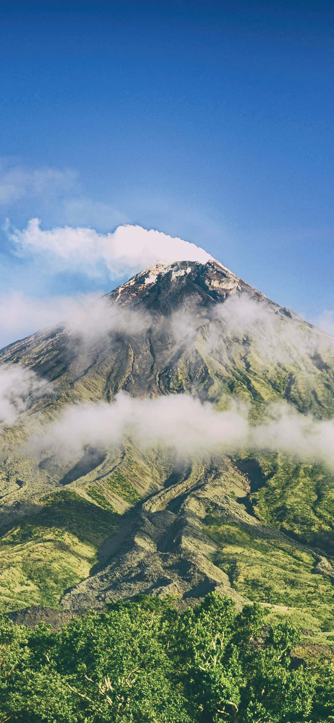 iPhone X Nature 桌布 壁紙 — 一座雄偉的綠色山峰，白雪覆蓋的山頂，在清澈的藍天下有旋轉的雲霧。