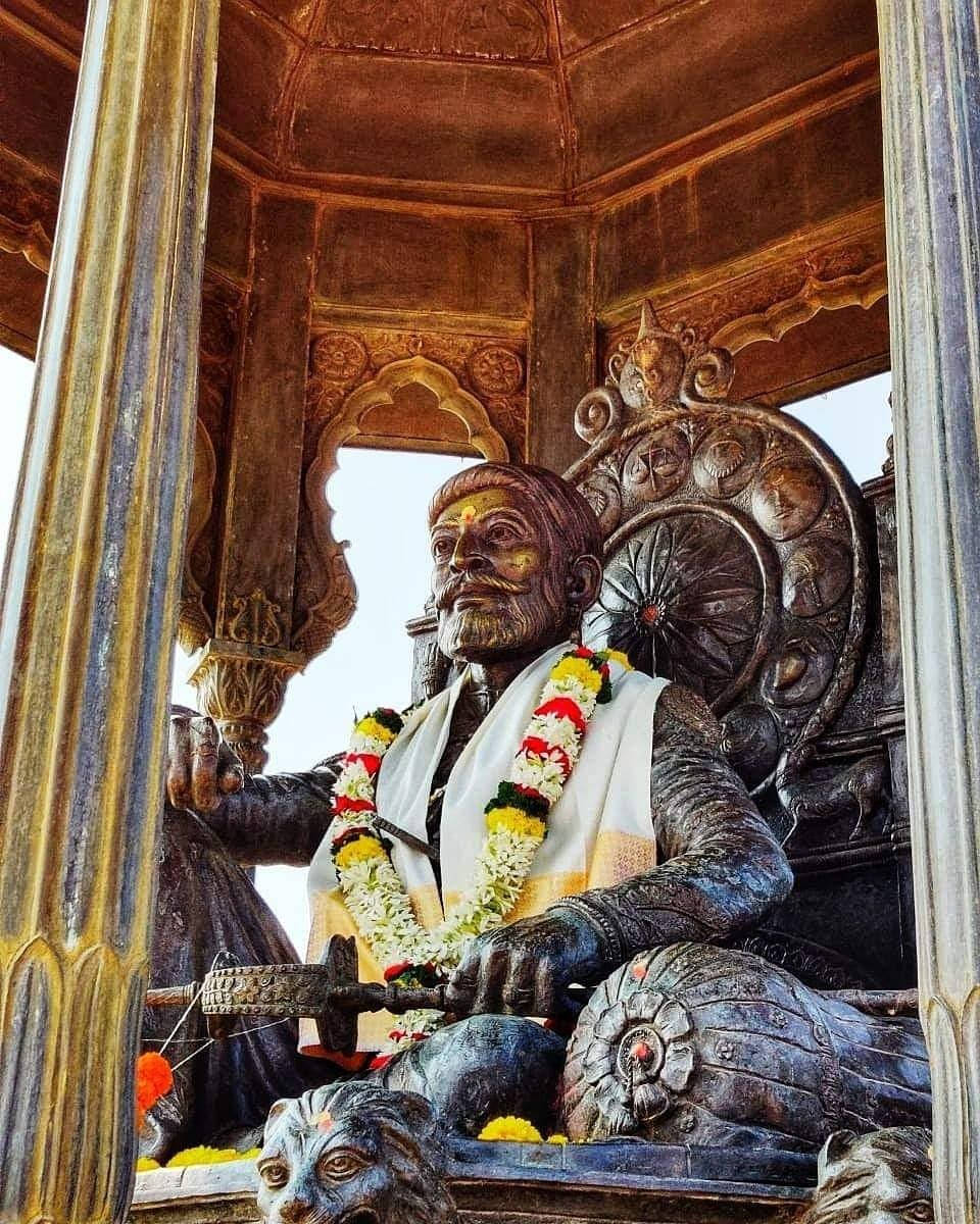 shivaji maharaj HD 桌布 壁紙 — 一座雄偉的銅像，一位鬍鬚男子坐在裝飾華麗的寶座上，寶座飾有花環，上方有雕刻華蓋。