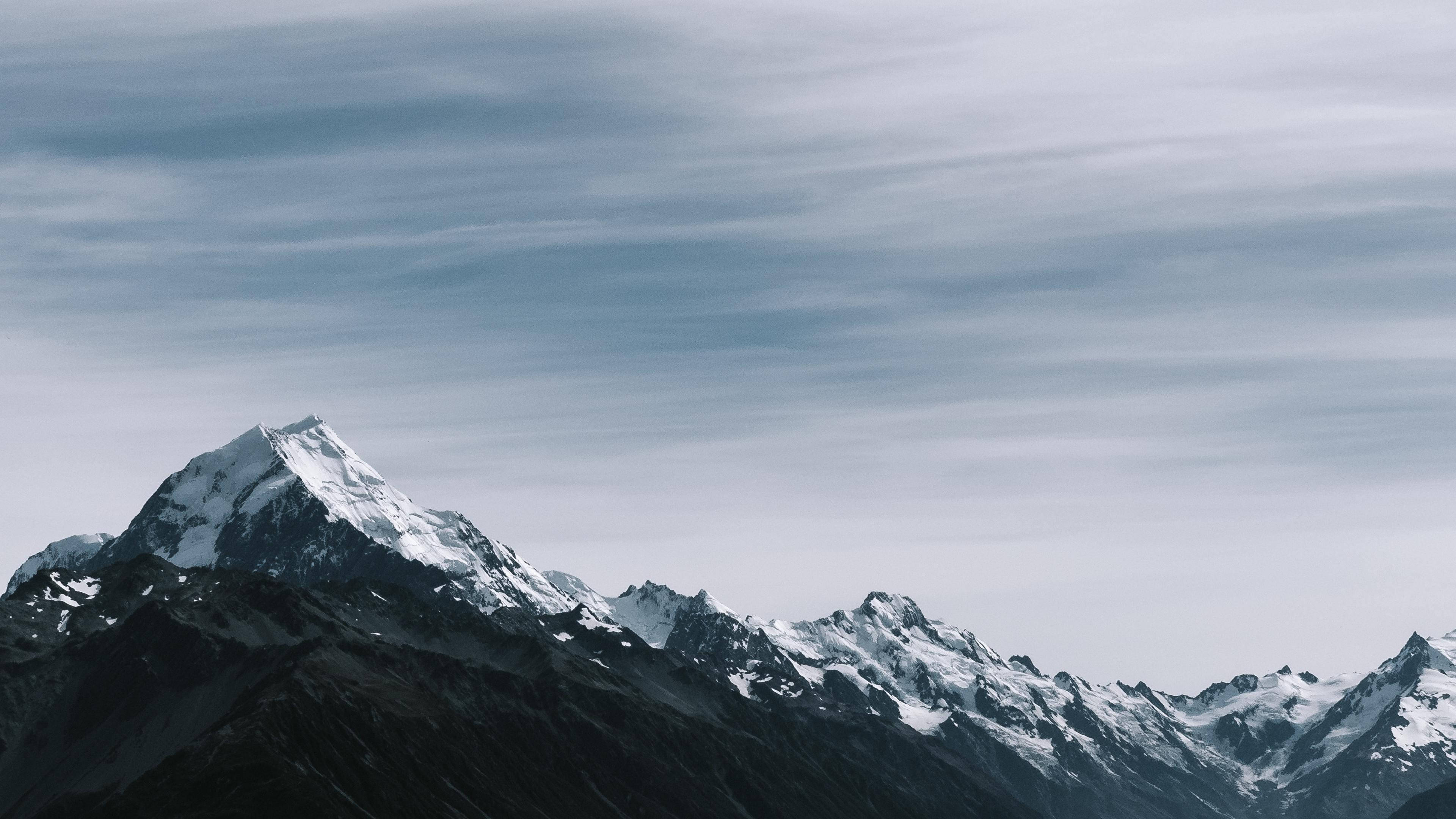 Mountain 桌布 壁紙 — 一座雄偉的雪山山峰在帶有條紋雲的柔和灰色天空下陡峭地聳立。