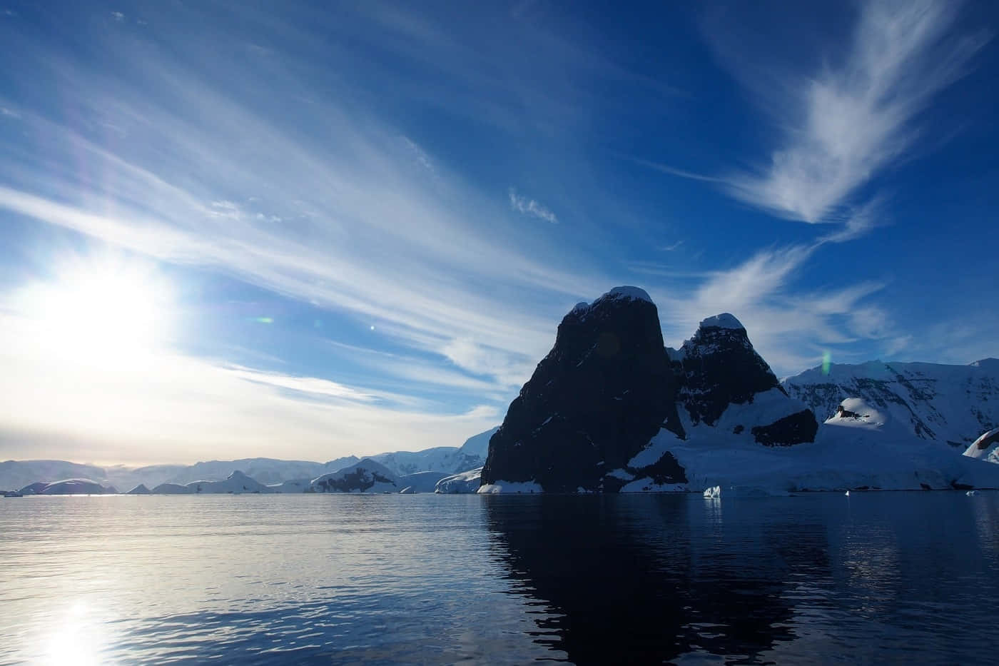 Antarctica 桌布 壁紙 — 一座雄偉的雪山從平靜的南極水域中升起，天空湛藍，點綴著縷縷薄雲。
